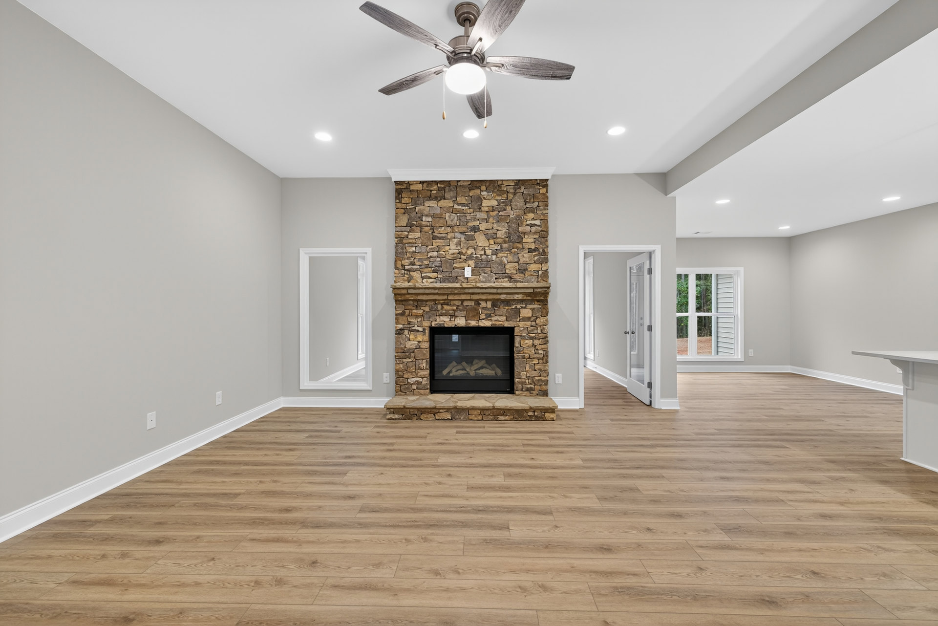 Living room with wood flooring, white walls, ceiling fan with light, glass-door fireplace stacked with firewood, white door and crown molding