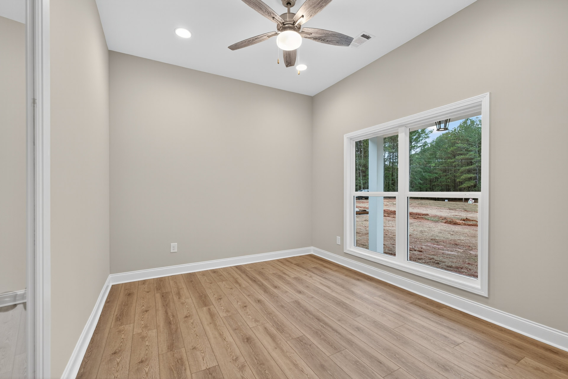 Ceiling fan with light fixture mounted on white plaster ceiling, wood laminate flooring, single window overlooking dirt field, neutral painted walls