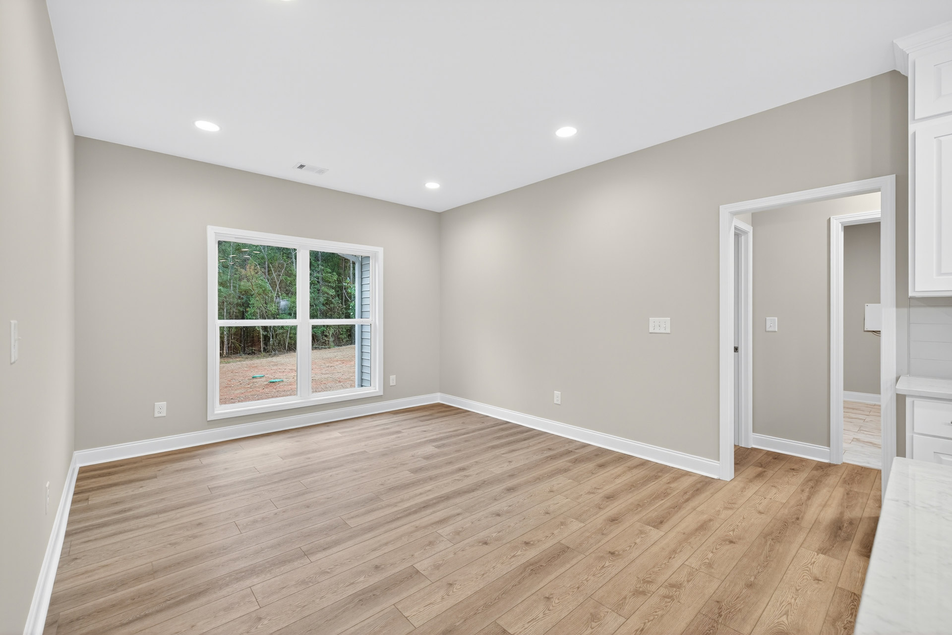 Room with hardwood flooring, large window overlooking yard, white ceiling with recessed lighting, white countertop visible in foreground
