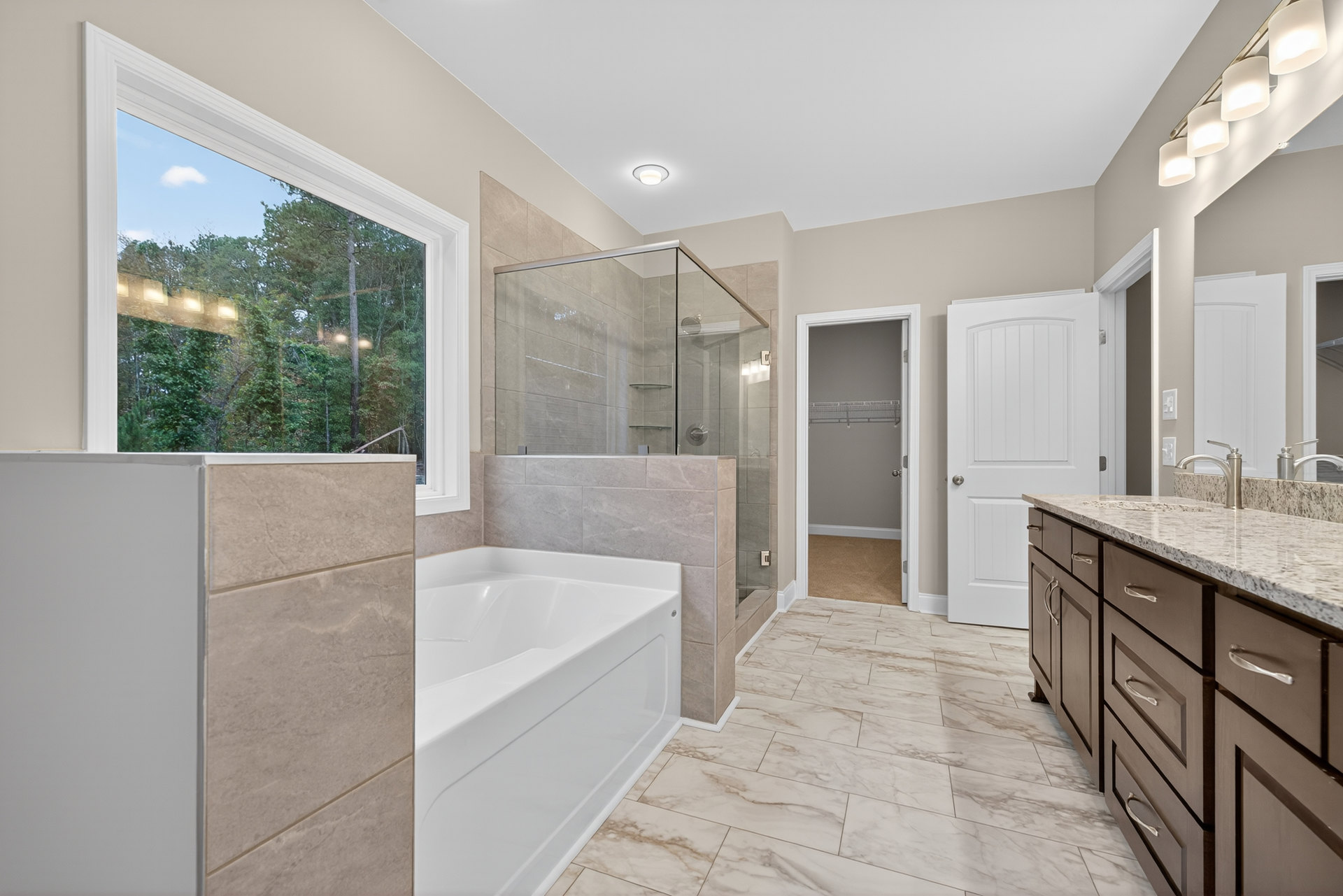 Bathroom featuring a white freestanding tub, glass-enclosed shower with tile walls, marble countertops, open closet door, four-light fixture, and window overlooking trees.