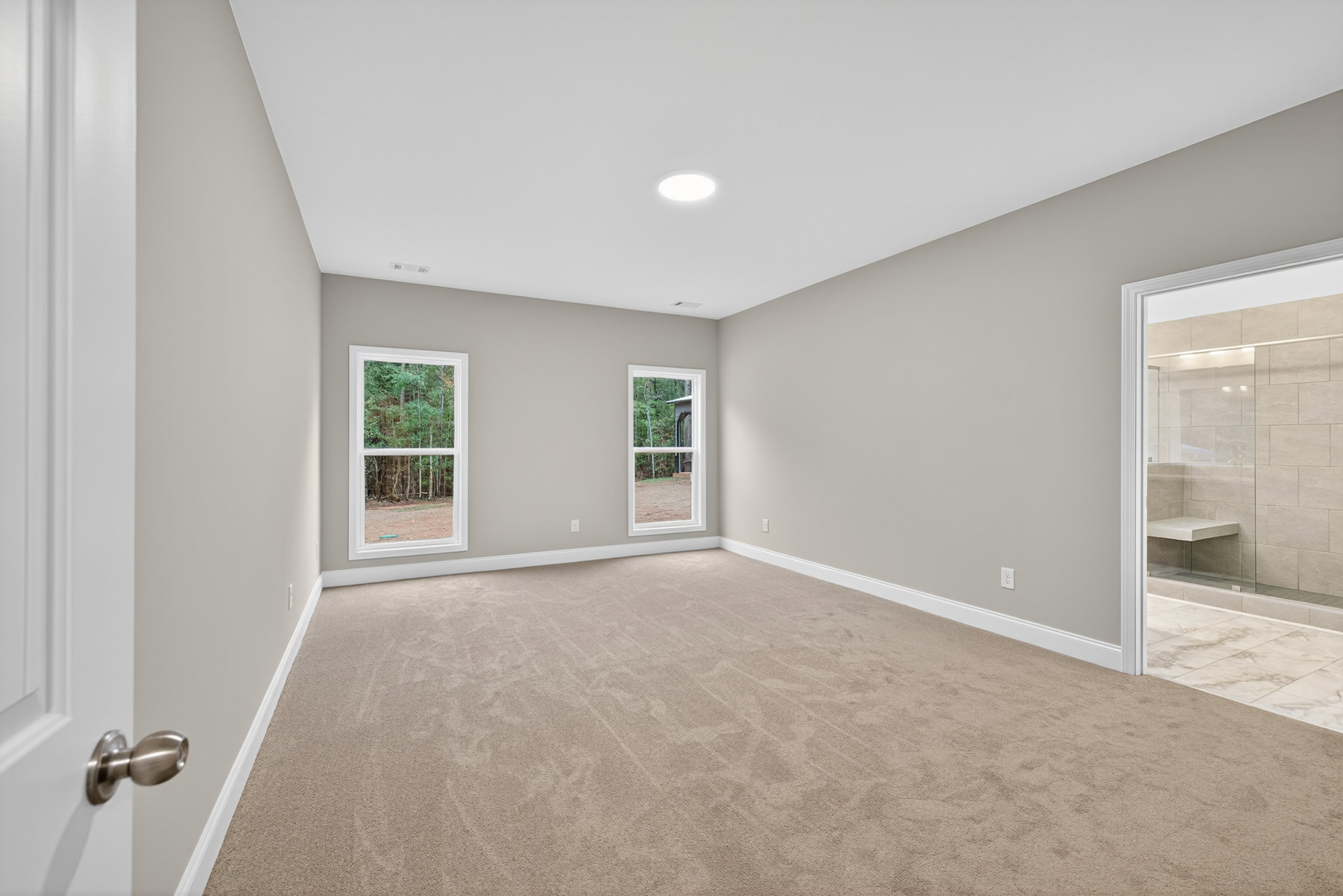 Beige carpeted room with large windows, white walls, ceiling light fixture, and views of trees and a neighboring house through the glass