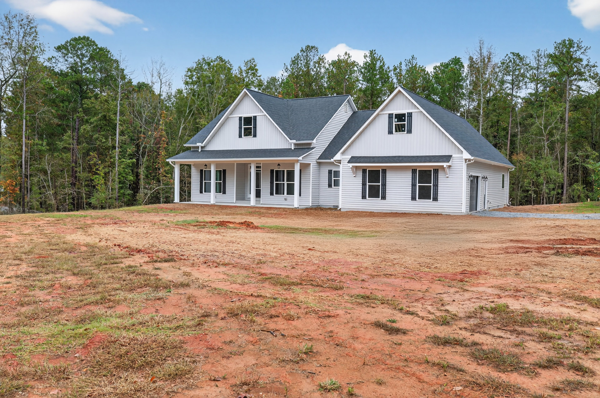Two-story house with black roof, white-framed windows, and large dirt yard bordered by trees under a cloudy sky