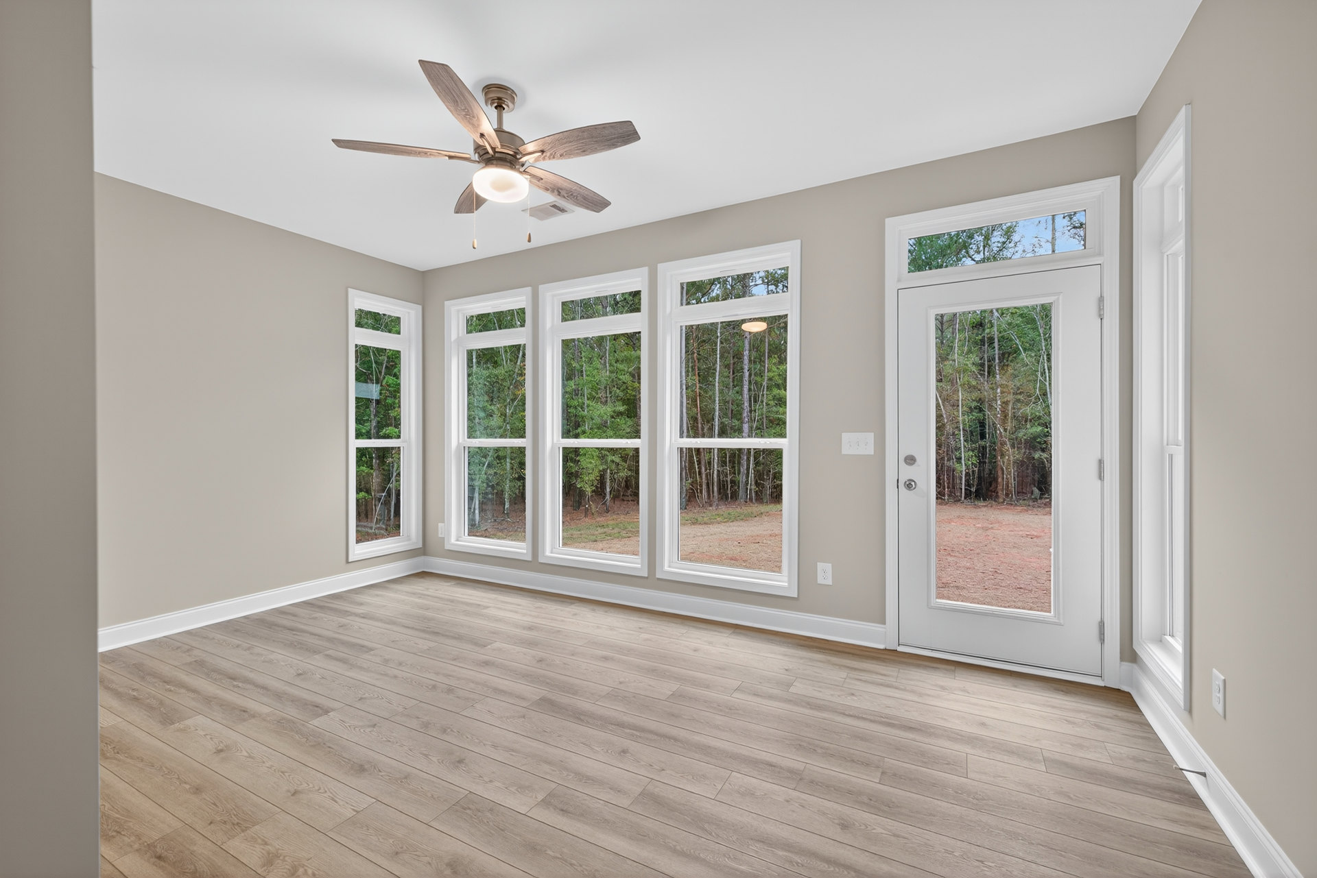 Wood flooring, white-framed windows overlooking trees, ceiling fan with light, door opening to forest view