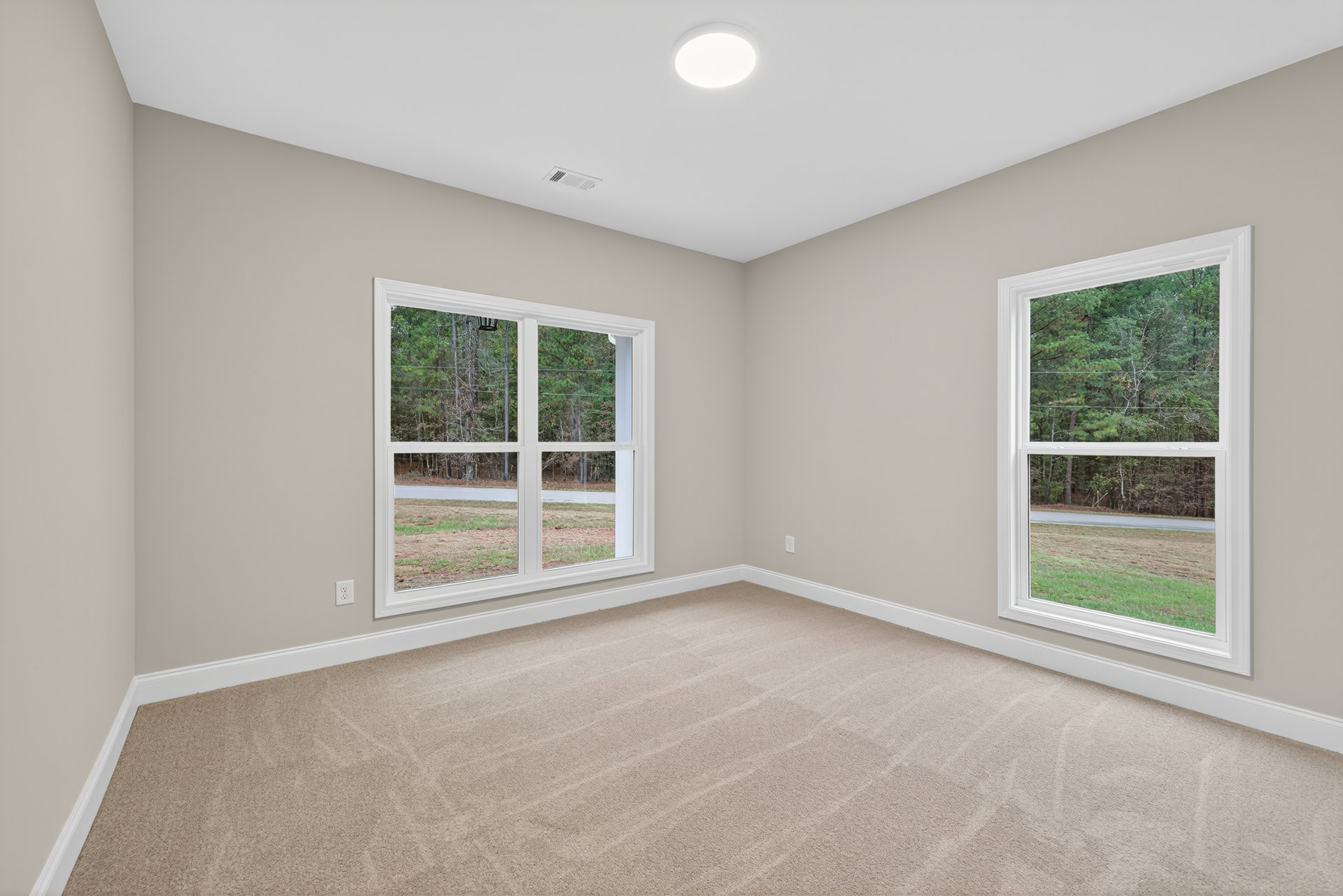 Carpeted bedroom with large window overlooking leafy trees, white walls, ceiling light fixture, and simple molding details