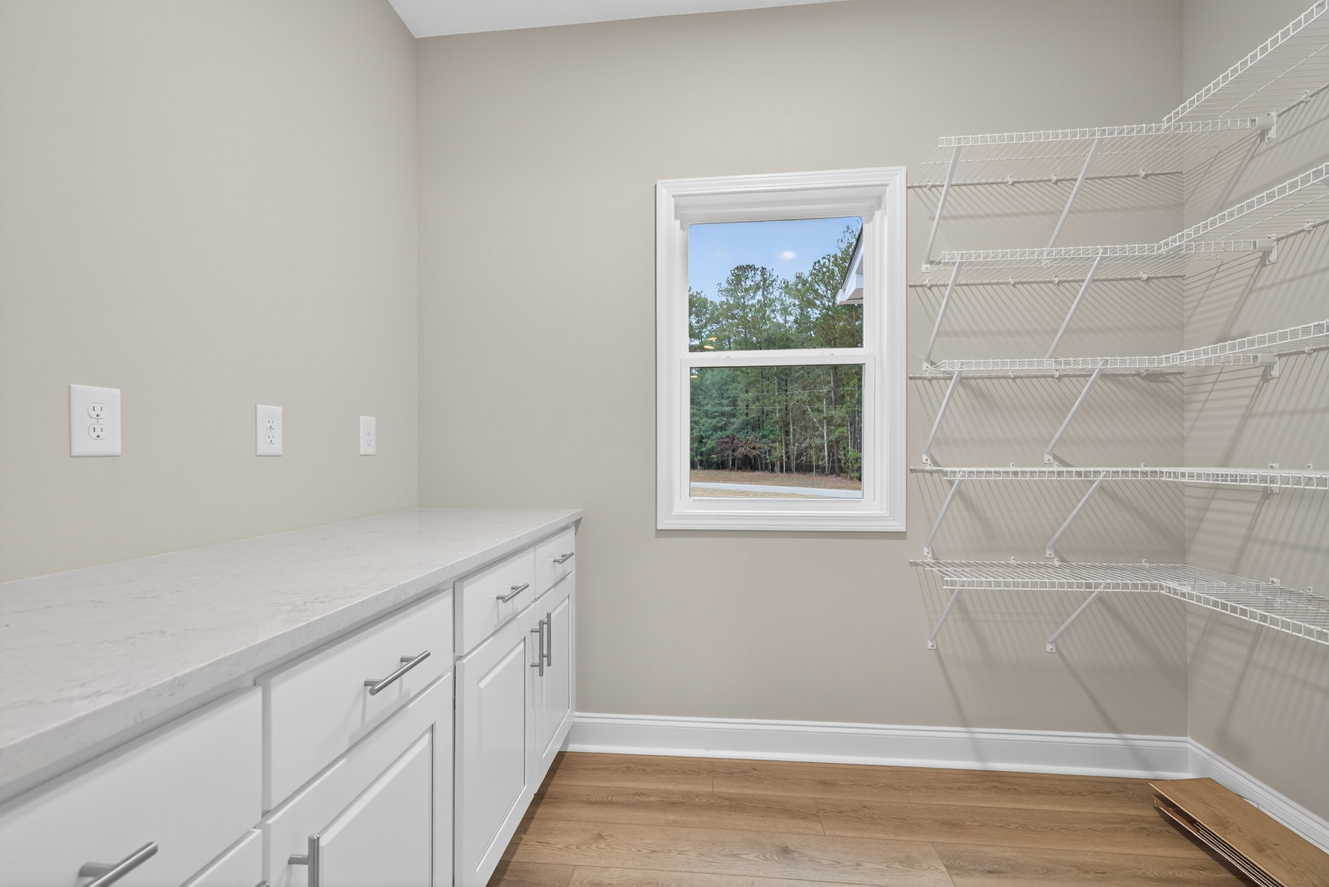 White cabinetry and open shelves beside a window with trees visible outside, wood flooring with white baseboard, white countertop with silver handles, stainless steel sink and