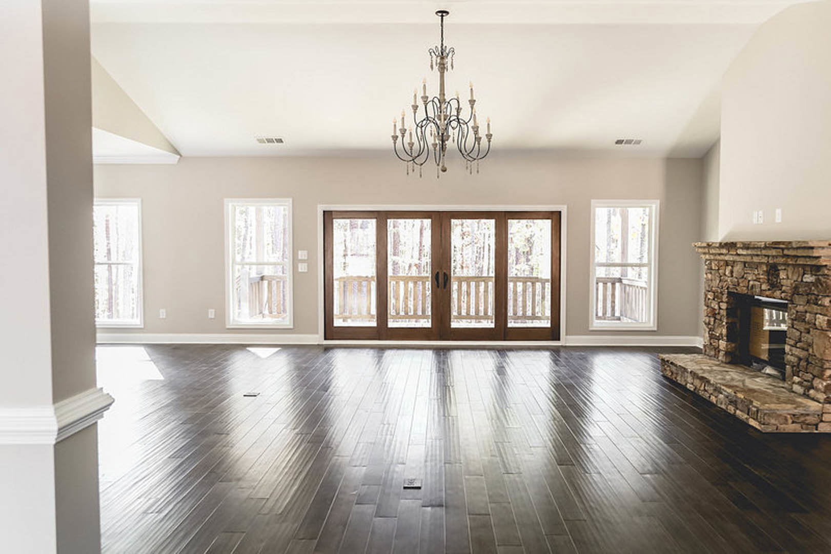 Spacious living room with hardwood flooring, ornate chandelier, glass-paneled double doors, fireplace with raised hearth, white wall molding, and large window
