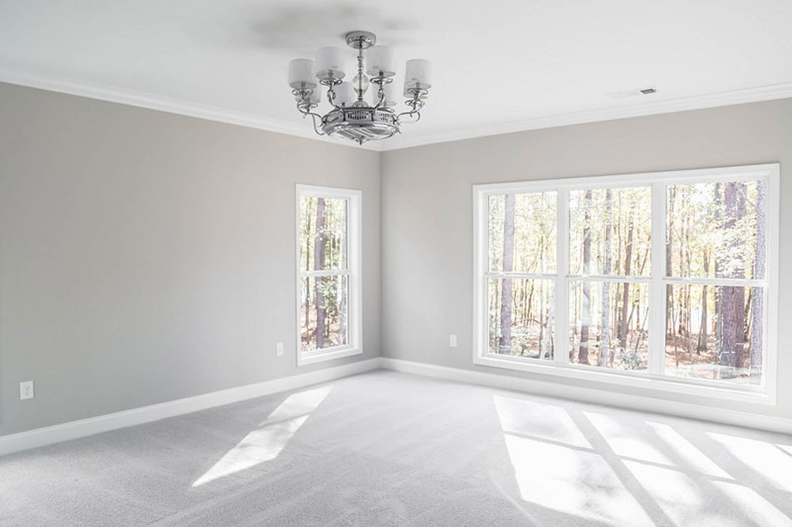 Chandelier hanging from white ceiling above plush white carpet, large windows with white trim framing views of green trees, smooth white walls with decorative molding