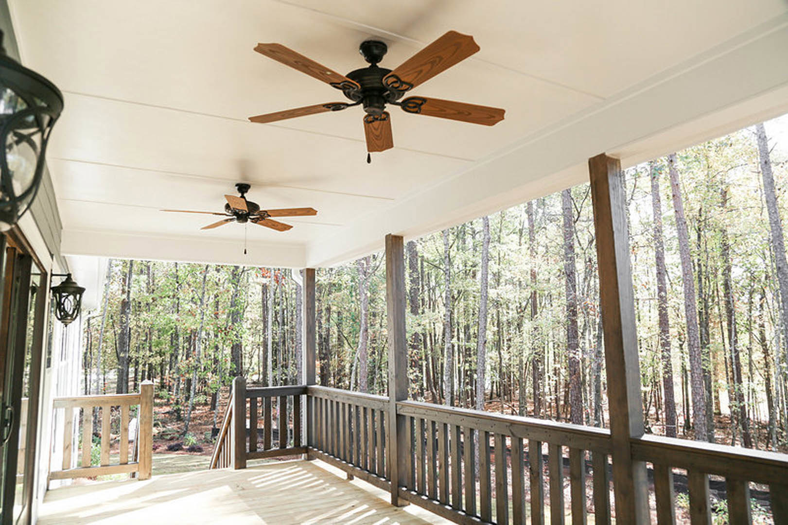 Ceiling fan with wood blades and integrated light mounted above a covered porch with wooden railing, surrounded by wooded landscape