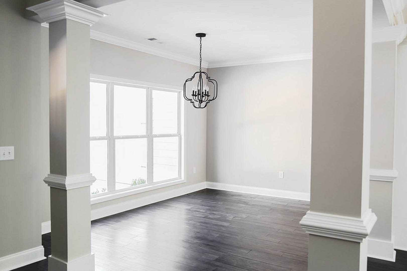 Black wood flooring with white trim, white pillar in foreground, white window and wall with black light switch and outlet, chandelier hanging from ceiling
