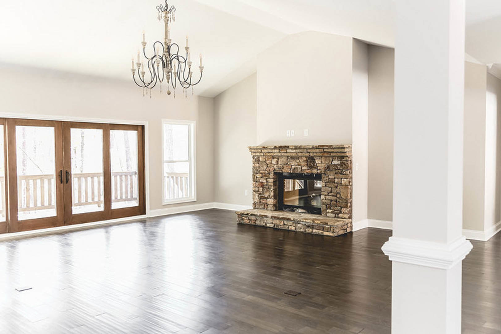 Living room featuring wood flooring, white molding, glass-paneled double doors, stone fireplace with glass door, candle-style chandelier, and large window