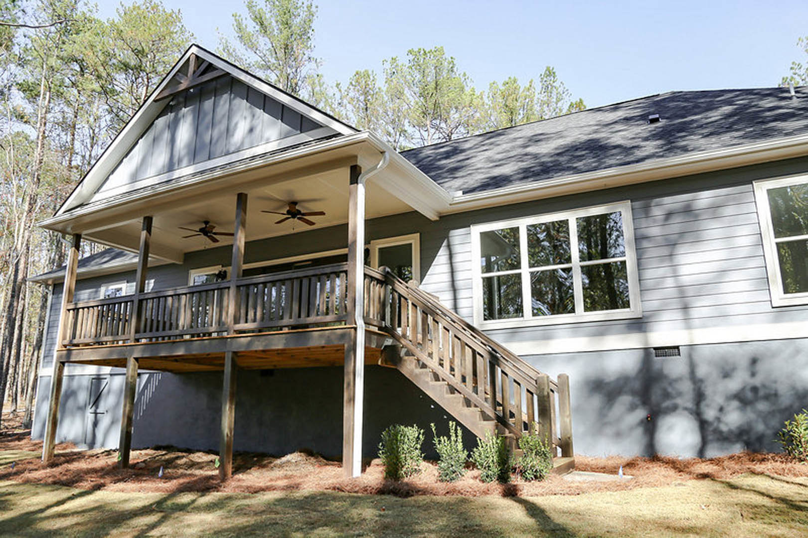 Two-story home with white siding, covered front porch, wood deck, large windows, ceiling fan, and landscaped plants and trees