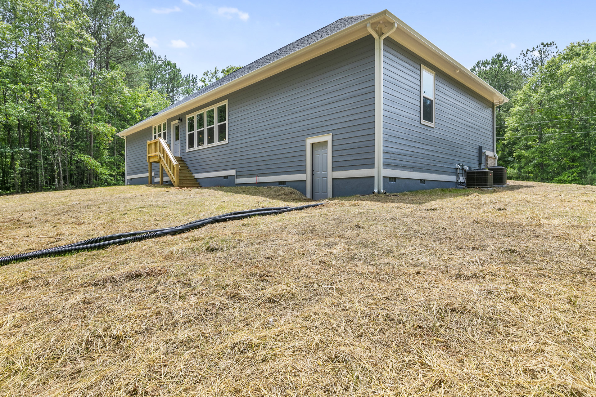 Two-story home with white siding, wooden front porch and stairs, black corrugated pipe along the lawn, mature trees in the background, large windows, and a close-up of the white