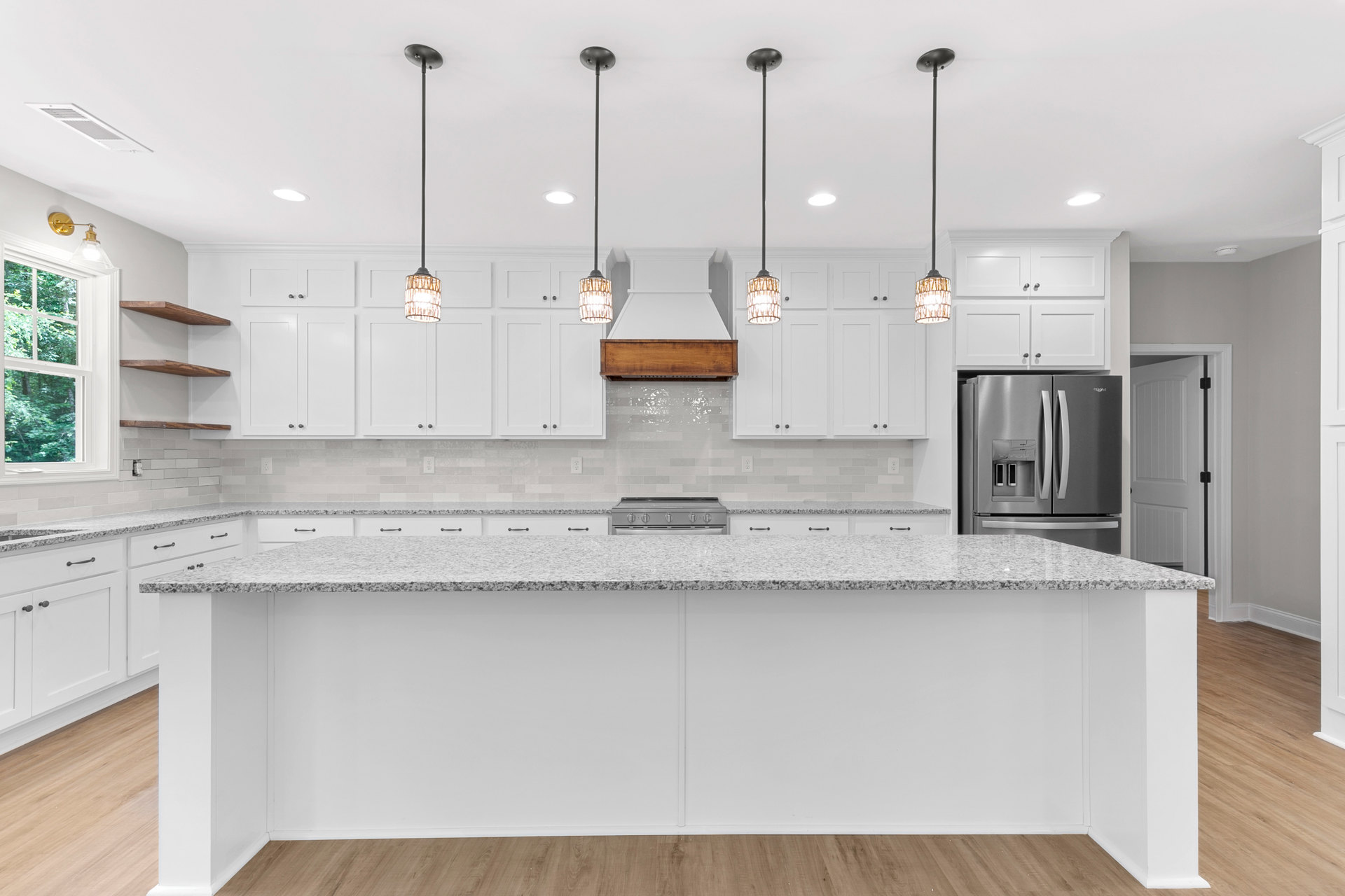 White kitchen with marble countertops, white cabinetry, stainless steel refrigerator, tile backsplash, window overlooking trees, wicker basket light fixture, and hardwood flooring.