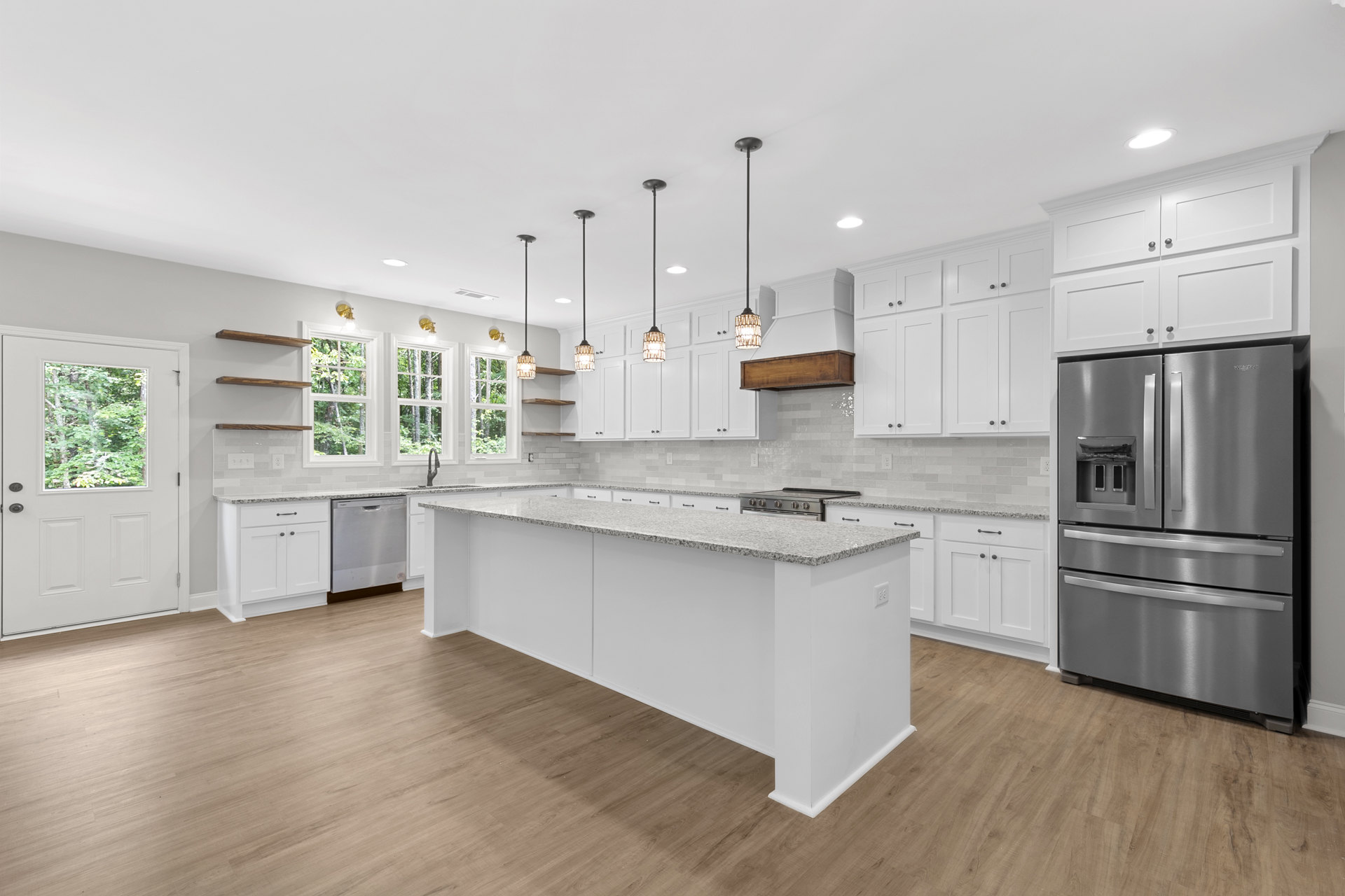Spacious kitchen featuring a large marble-topped island, stainless steel refrigerator with double drawers, white cabinetry, a window overlooking trees, and a white door with glass