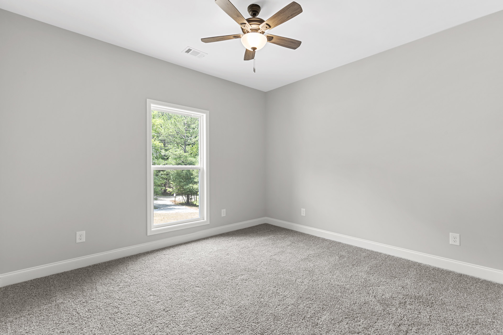 Neutral-toned bedroom with carpet flooring, white walls, ceiling fan with light fixture, single window showing leafy trees outside