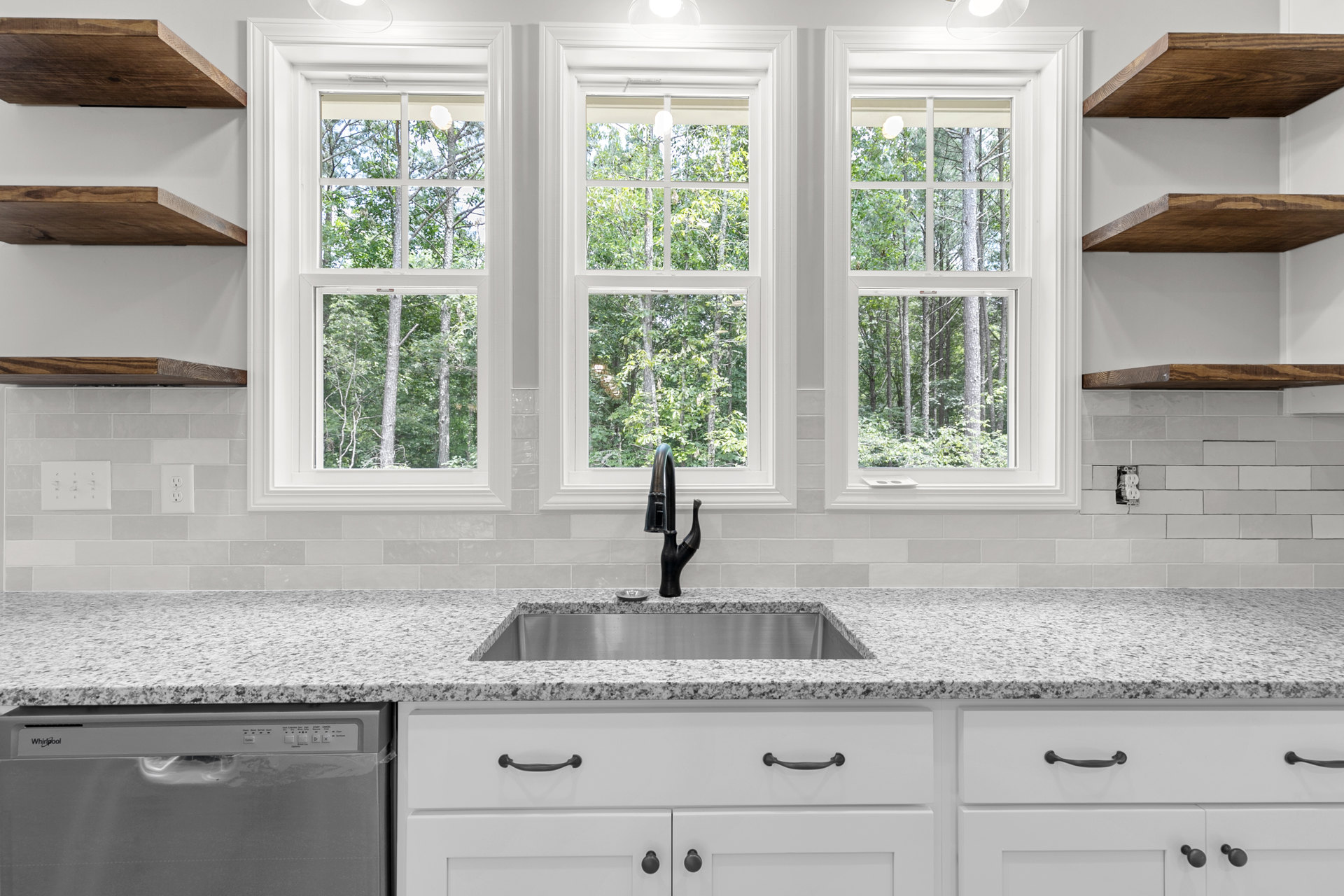 White kitchen with shaker cabinets, quartz countertops, stainless steel dishwasher, undermount sink and chrome faucet, large window overlooking trees, white trim and drawers