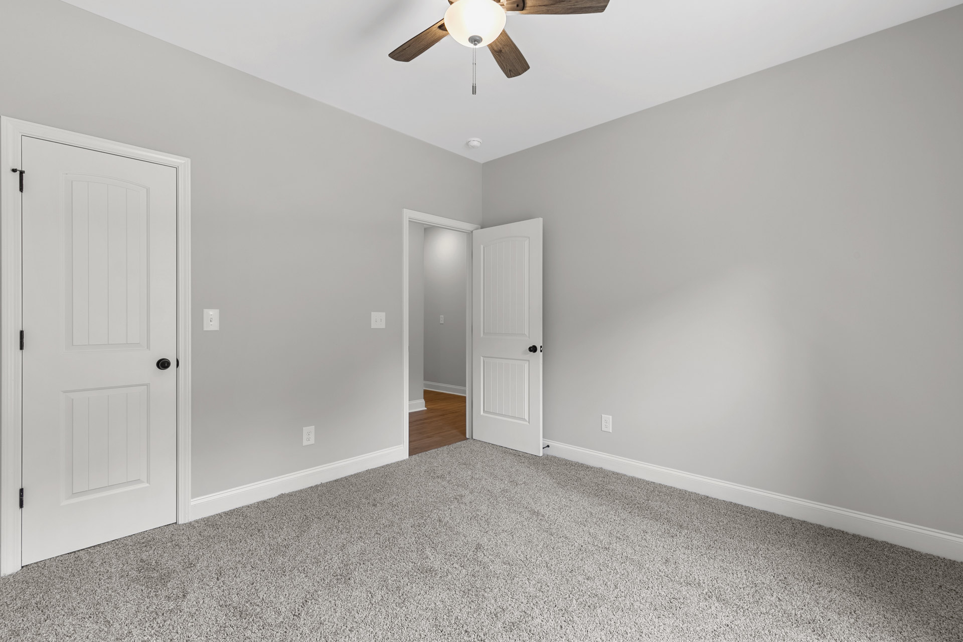 Wood floor room with white paneled door featuring black knob, ceiling fan with light fixture, white walls, and plaster ceiling.