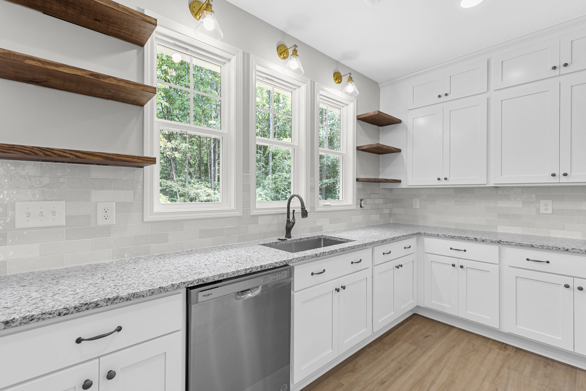 White kitchen cabinets, stainless steel dishwasher, granite countertop, chrome faucet, window with view of trees, white electrical outlet on backsplash
