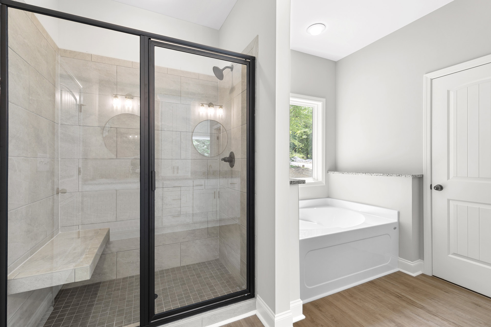 Bathroom with glass shower enclosure, built-in bench, circular wall mirror, freestanding white bathtub, white door with black handle, tile flooring, and modern light fixture