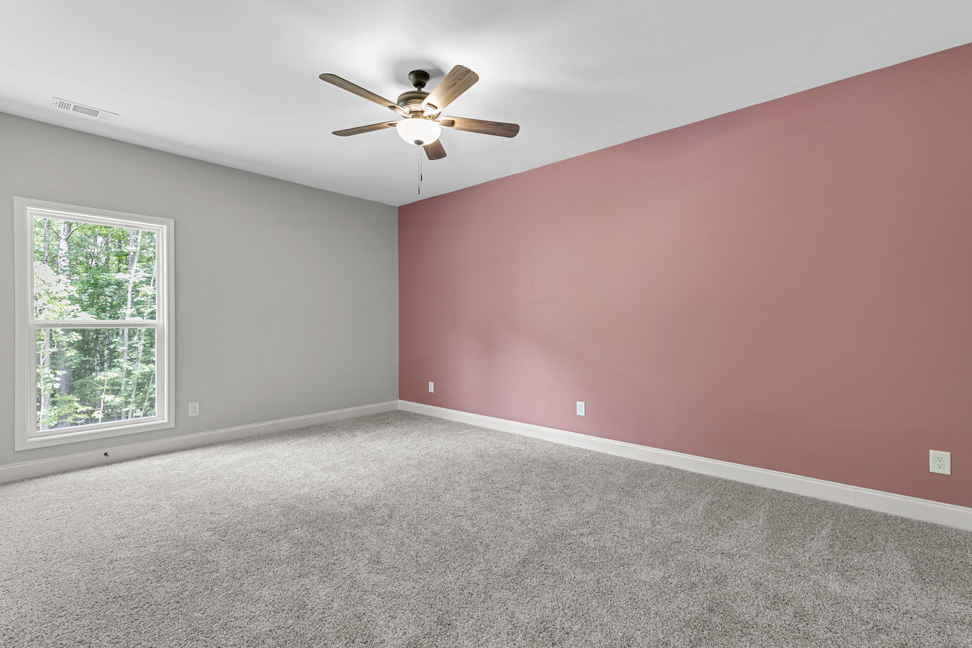 Pink-walled room with carpet flooring, ceiling fan with light fixture, and window overlooking trees.