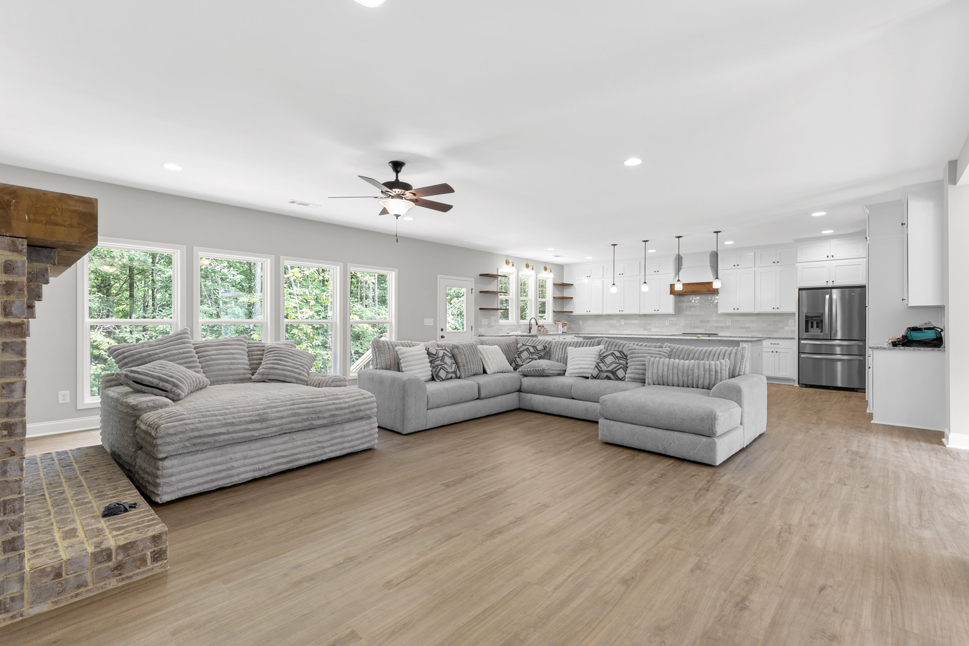 Spacious living room featuring a large sectional couch with multiple pillows, wood flooring, exposed brick wall with white outlet, and a pile of towels beside a brick fireplace