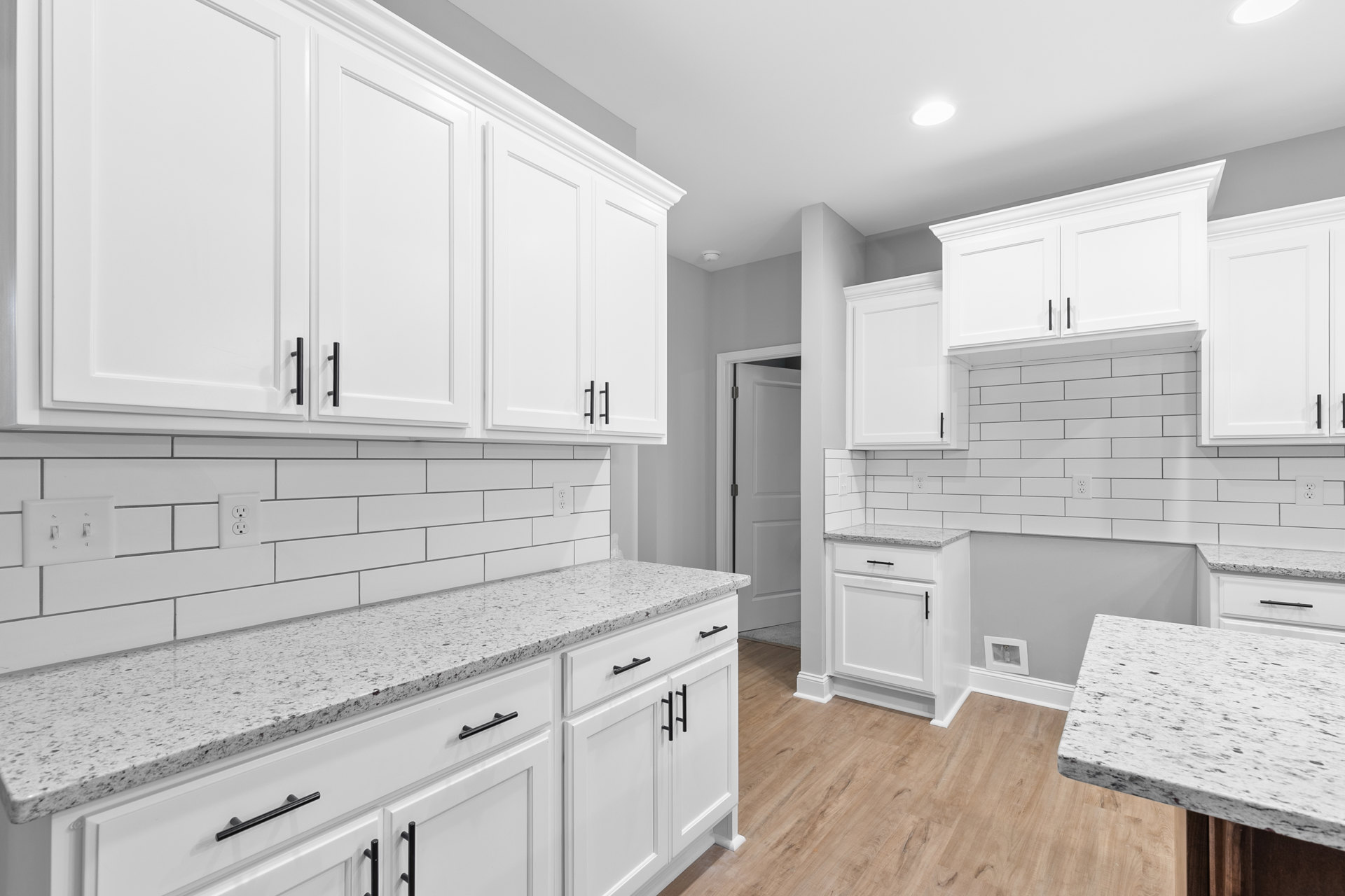 White kitchen with marble countertops, white cabinets featuring black handles, stainless steel sink, and built-in appliances; white tile backsplash and recessed lighting.