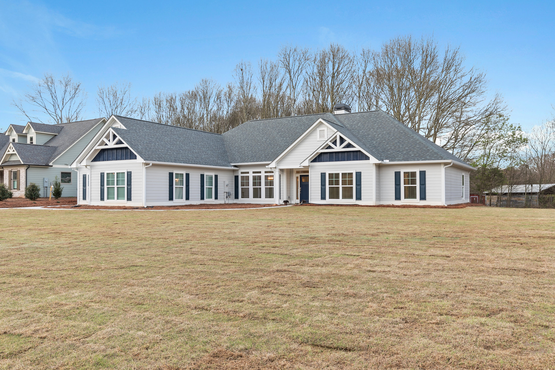 White farmhouse-style home with blue front door, multiple windows with blinds, expansive green lawn, mature trees, and clear sky in the background