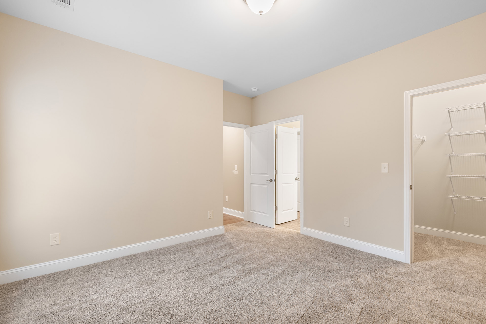 Beige carpeted room with white closet shelves, white door featuring silver handle, ceiling light fixture, and simple wall molding