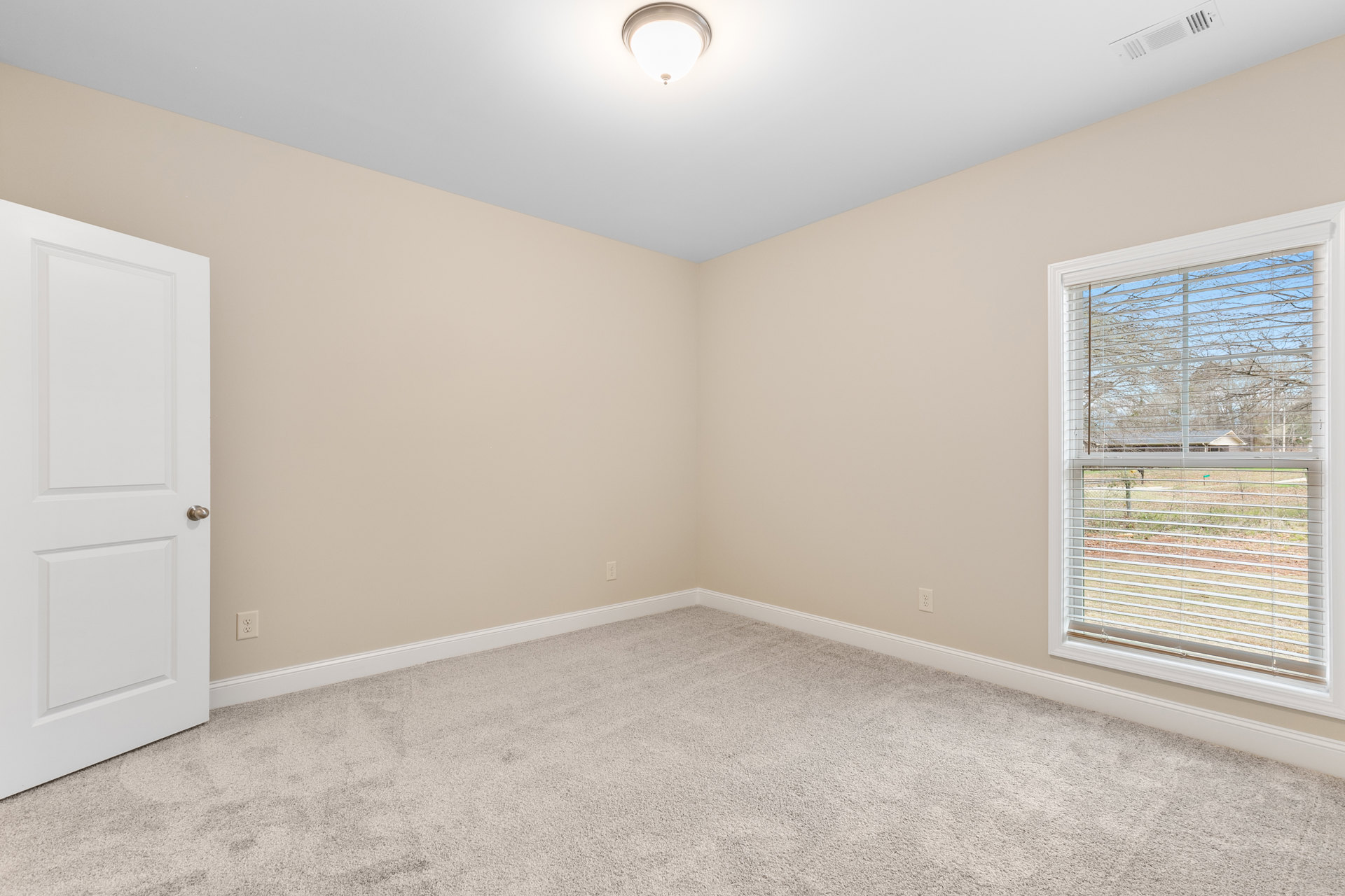 Carpeted room with white walls, window fitted with blinds, white door featuring a silver handle, ceiling-mounted light fixture, and simple molding along the baseboards.