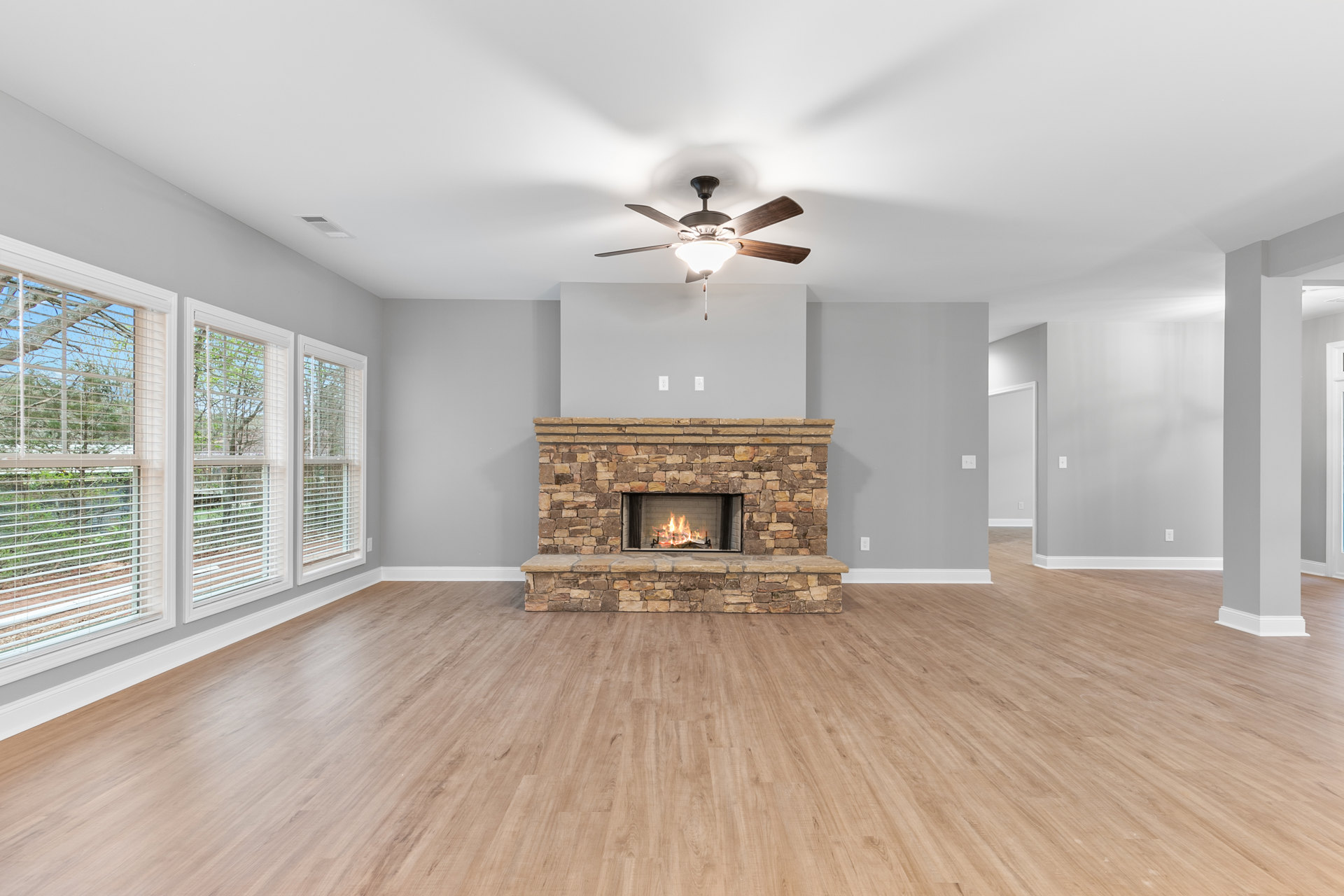 Living room with hardwood floors, stone fireplace with burning fire, ceiling fan and light fixture, window with white blinds, crown molding along ceiling