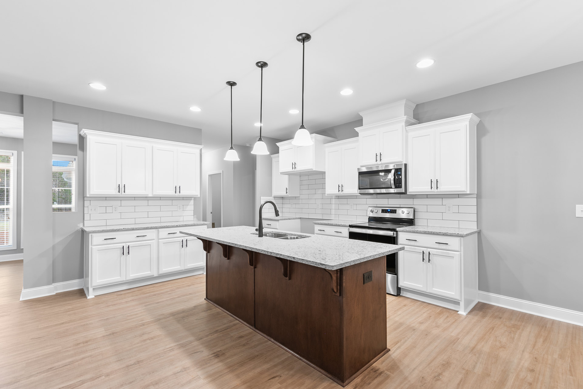 White kitchen with shaker cabinets, central island featuring built-in sink, stainless steel microwave, light stone countertops, wood flooring, and brushed metal hardware.