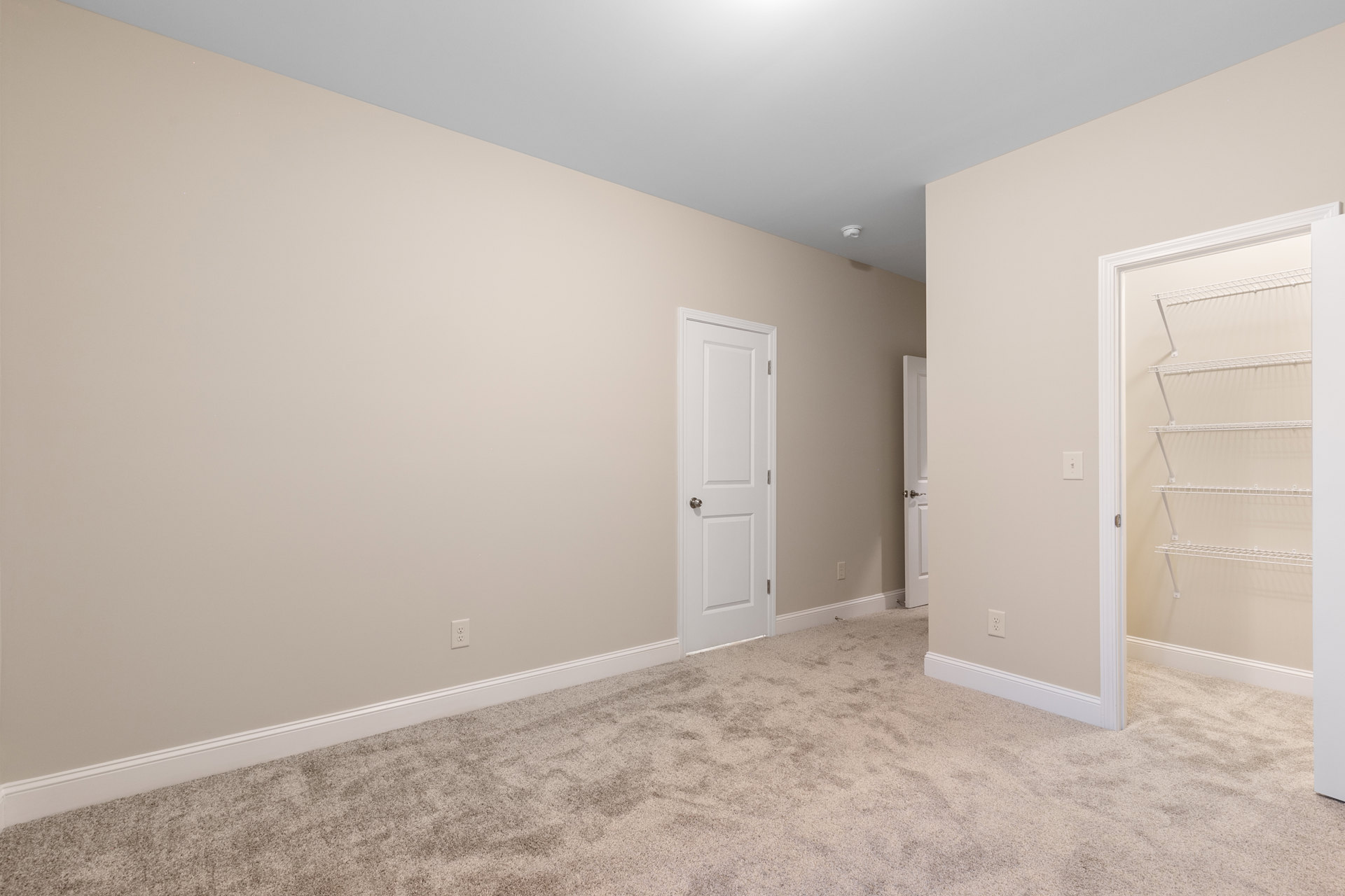 Beige-walled room with white doors, silver door knob, white wall shelf, carpeted floor, and hallway featuring two doors