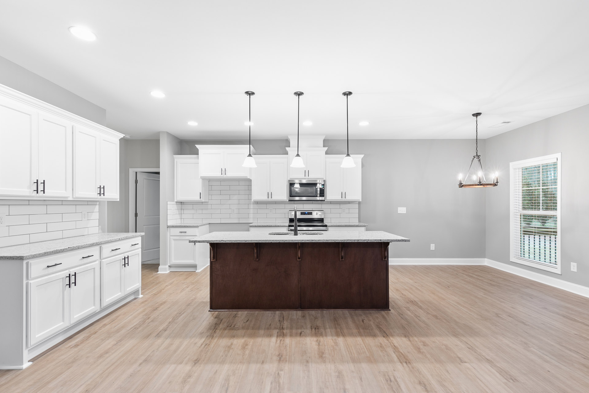 White kitchen cabinets, large marble-topped island, stainless microwave with interior light, sink set in white countertop, window with blinds, wood flooring, brown door