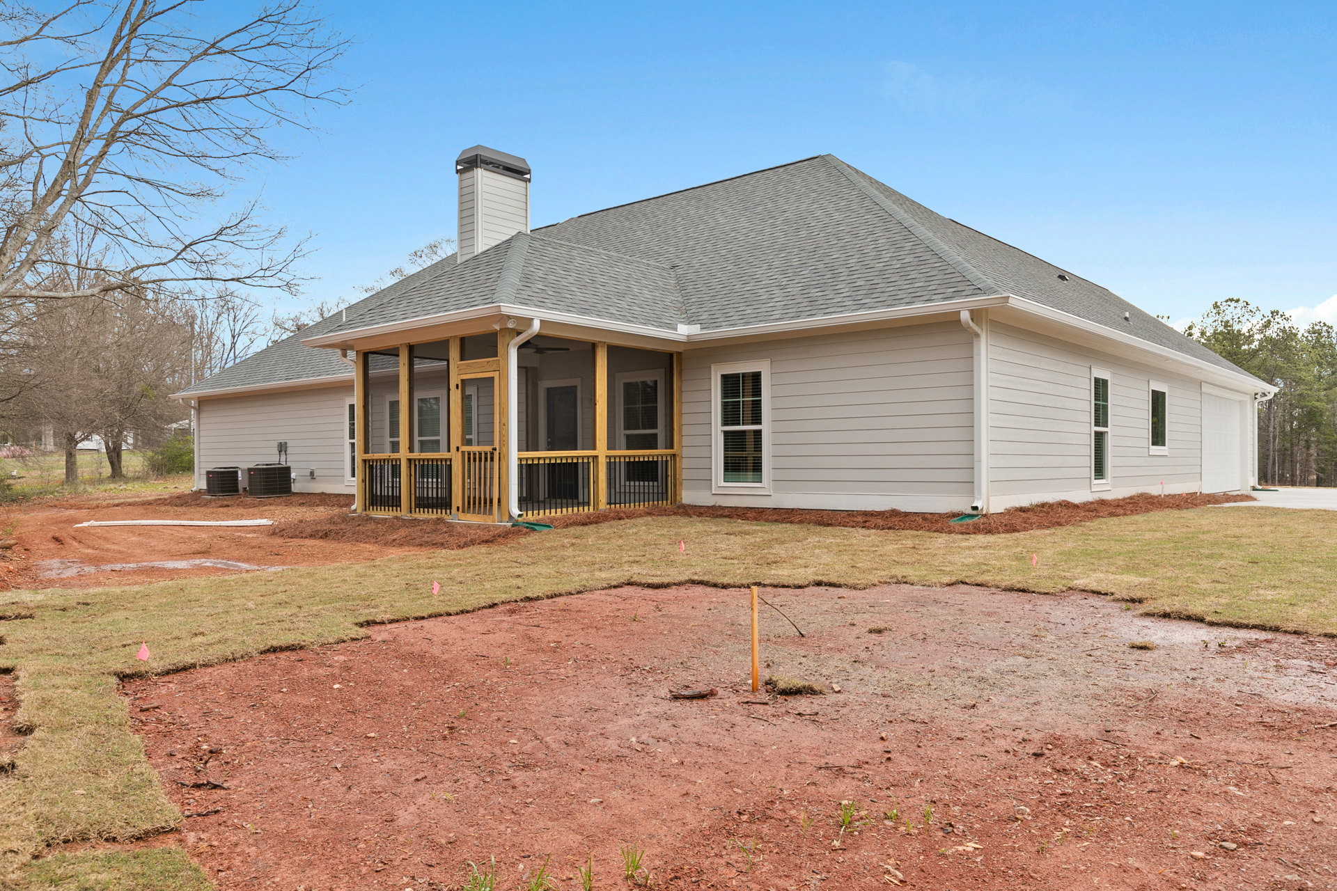 Two-story house with white-framed windows, wood porch and railing, bare tree, dirt patch in front yard, light-colored siding, and open grassy lot.
