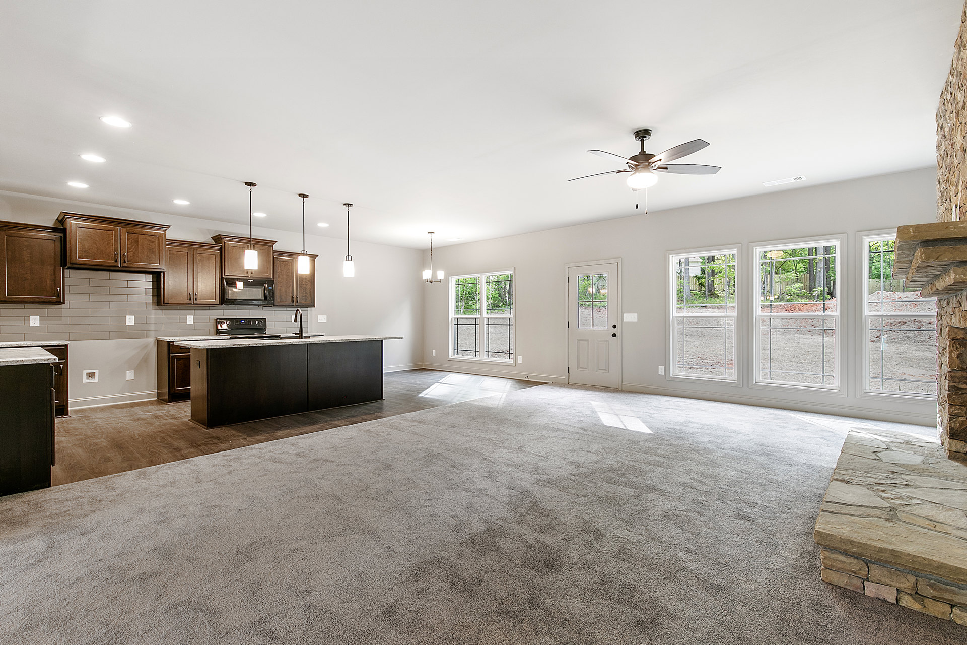 Open concept living room and kitchen featuring a ceiling fan with light, stone accent wall and ledge, kitchen counter with sink, cabinetry, microwave, and window overlooking trees.