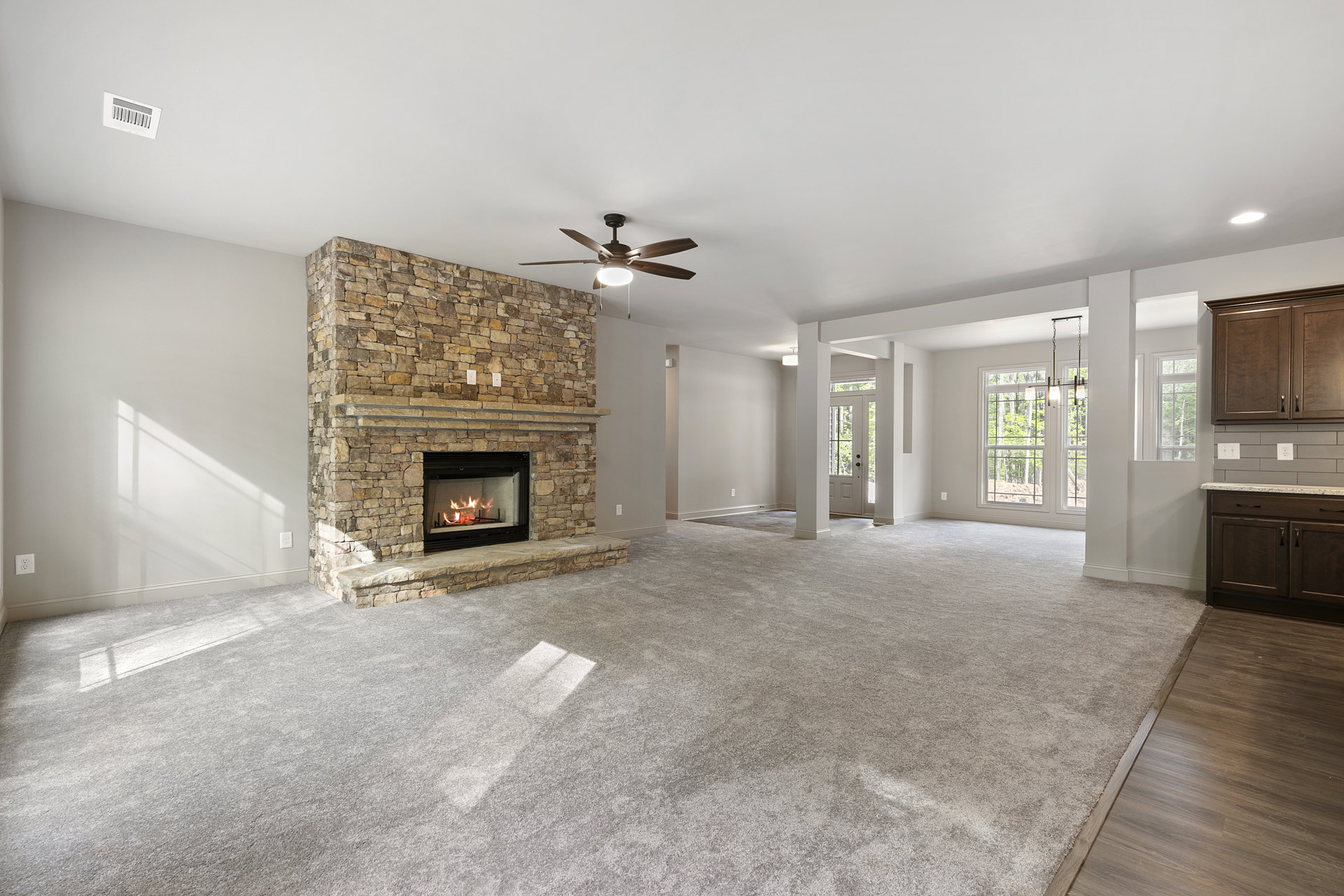 Living room with carpeted floor, stone fireplace with burning fire, ceiling fan with light fixture, white wall vent, wood cabinetry, neutral walls