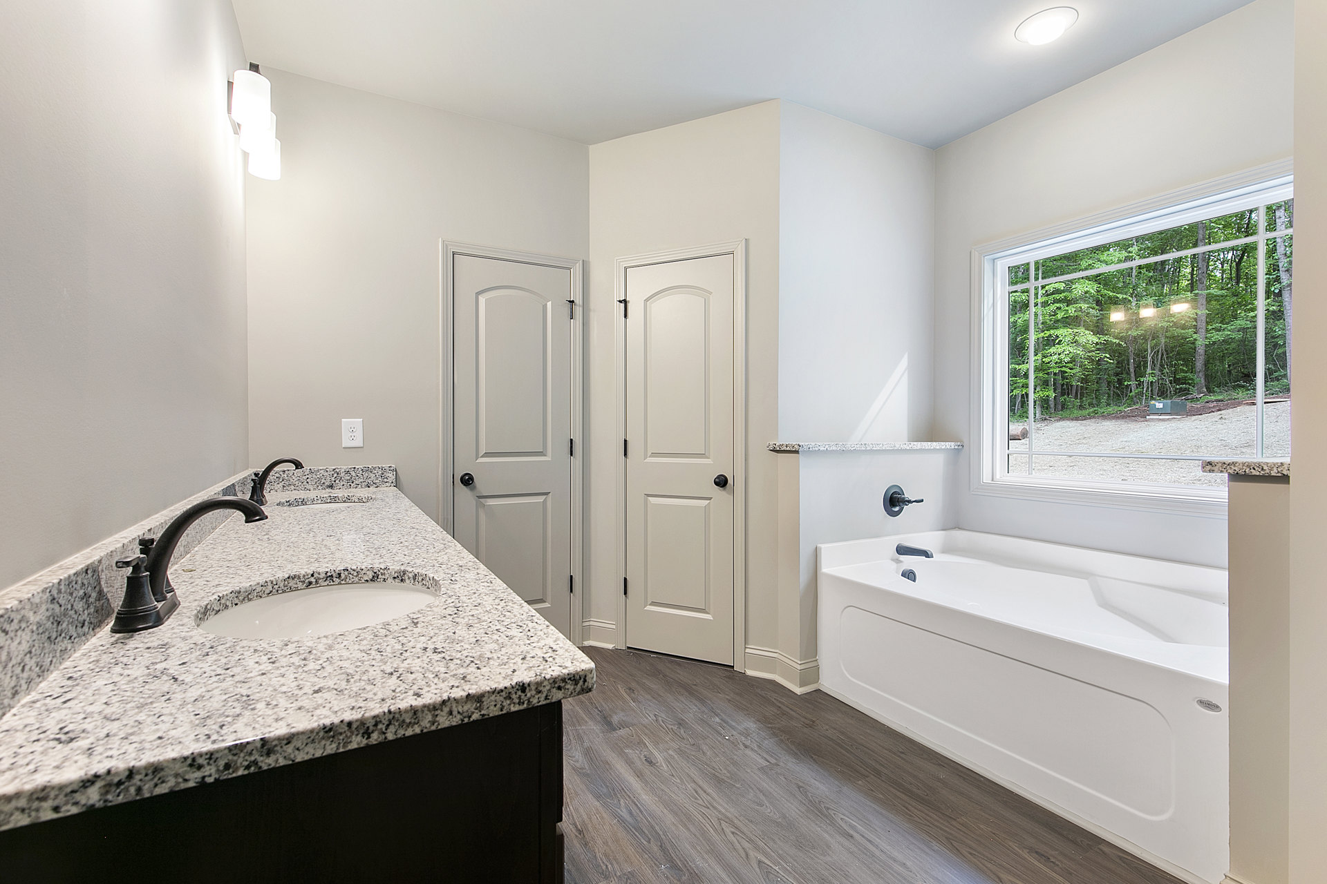 Bathroom featuring a marble countertop vanity with chrome faucet, freestanding white bathtub on wood flooring, white door with black knob, and window overlooking trees.
