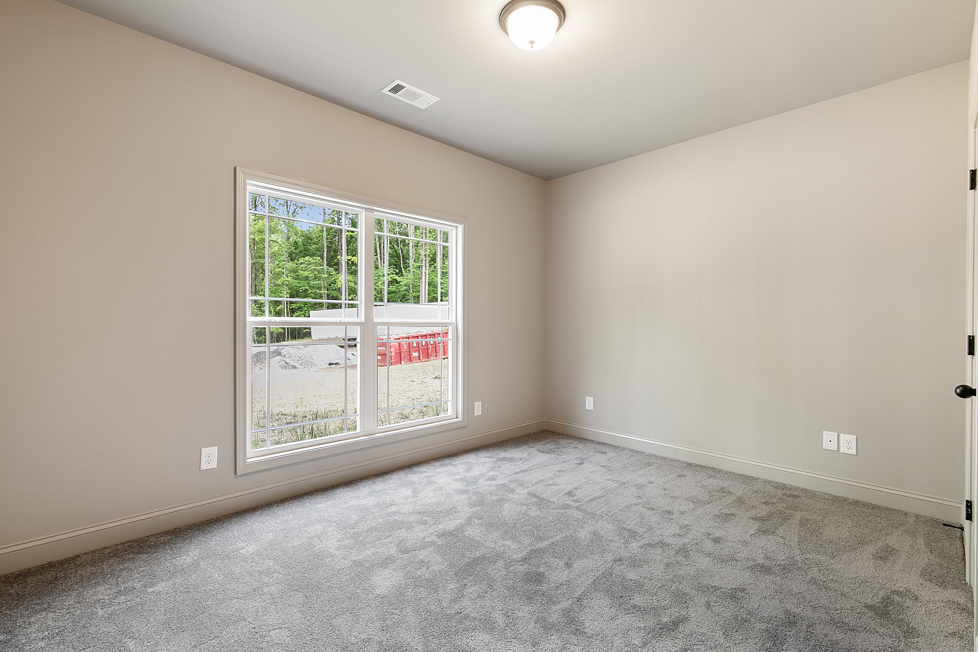 Grey carpeted room with white walls, large window showing trees and dirt outside, ceiling vent, round ceiling light fixture, and red container with white lettering.