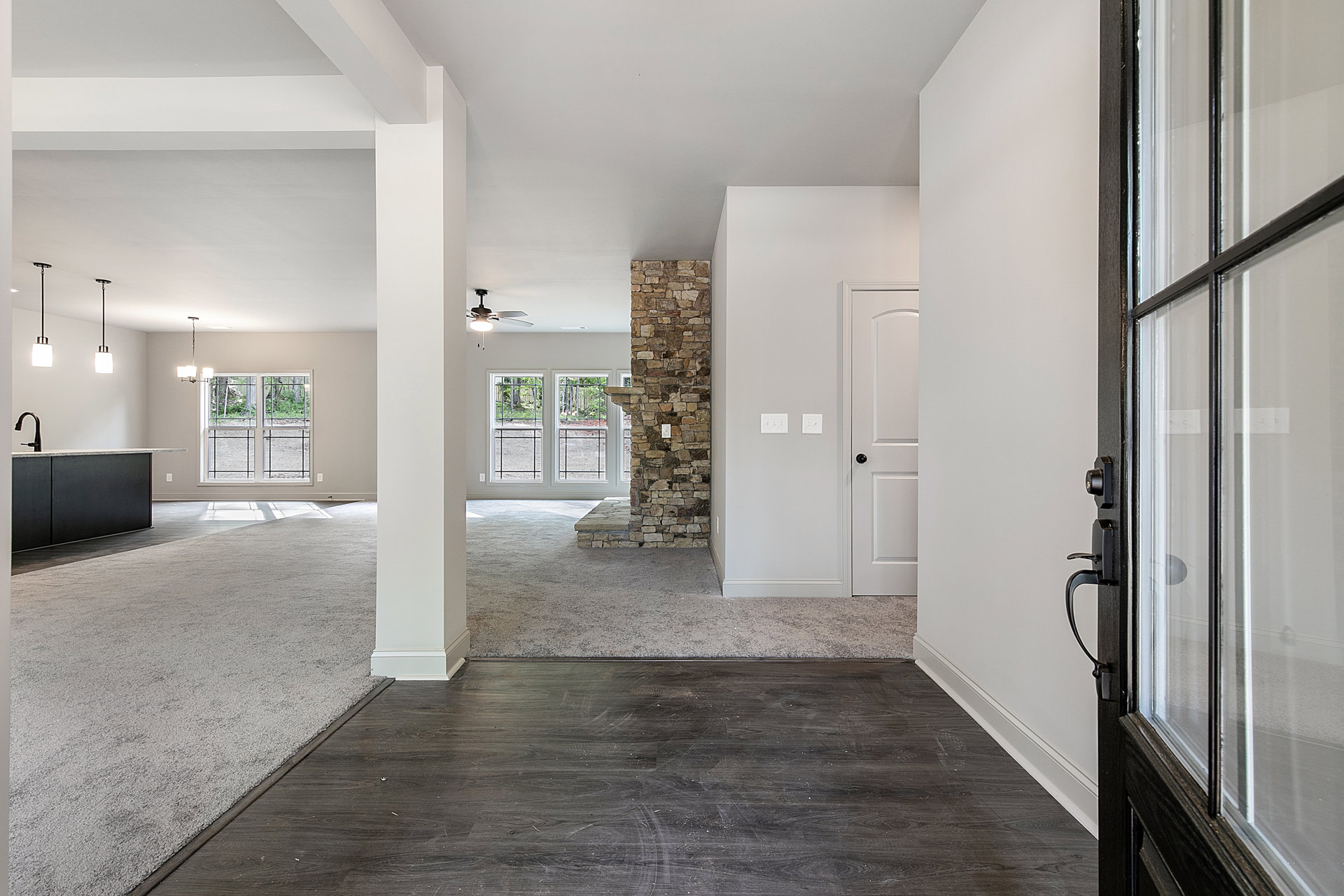 Black wood floor with white spots, white carpet, stone fireplace wall with light switch, large window overlooking forest, close-up of wooden door.
