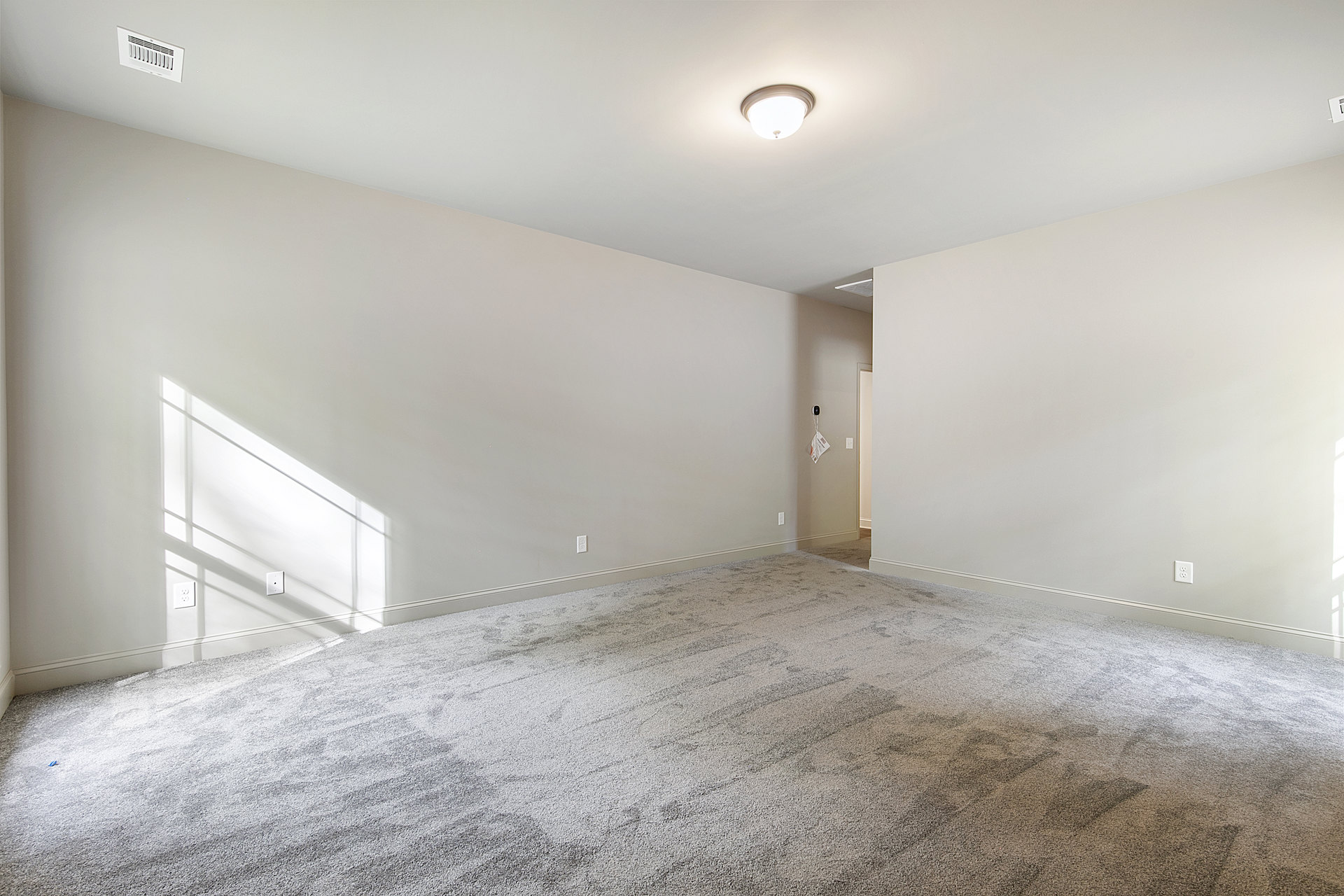 White plaster walls with soft shadows, ceiling-mounted light fixture, white air vent, beige carpet flooring, doorway visible in corner