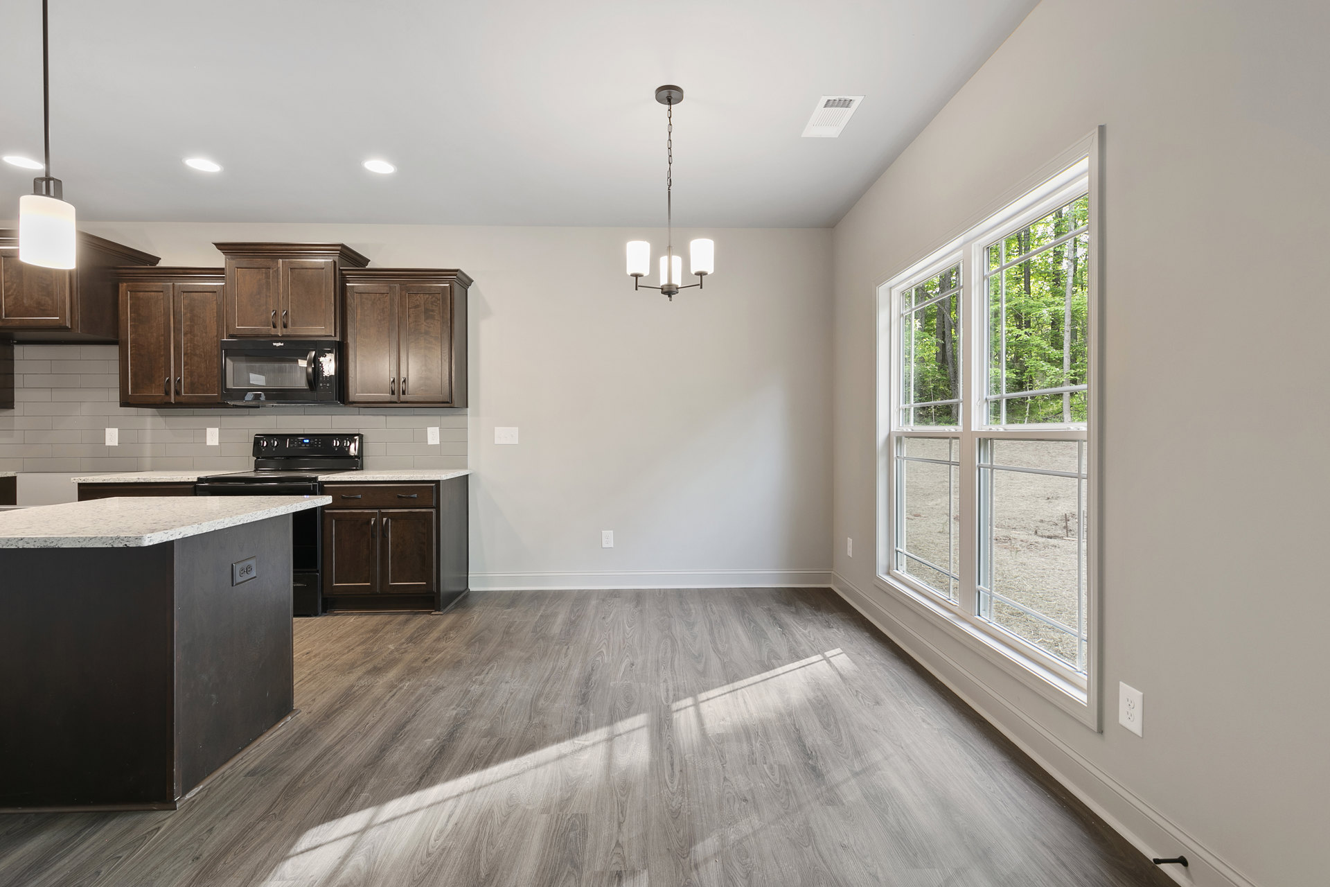 Open kitchen and dining area featuring wood floors, white walls, black cabinetry, marble-topped island, built-in black microwave with handle, window overlooking trees, and close-up