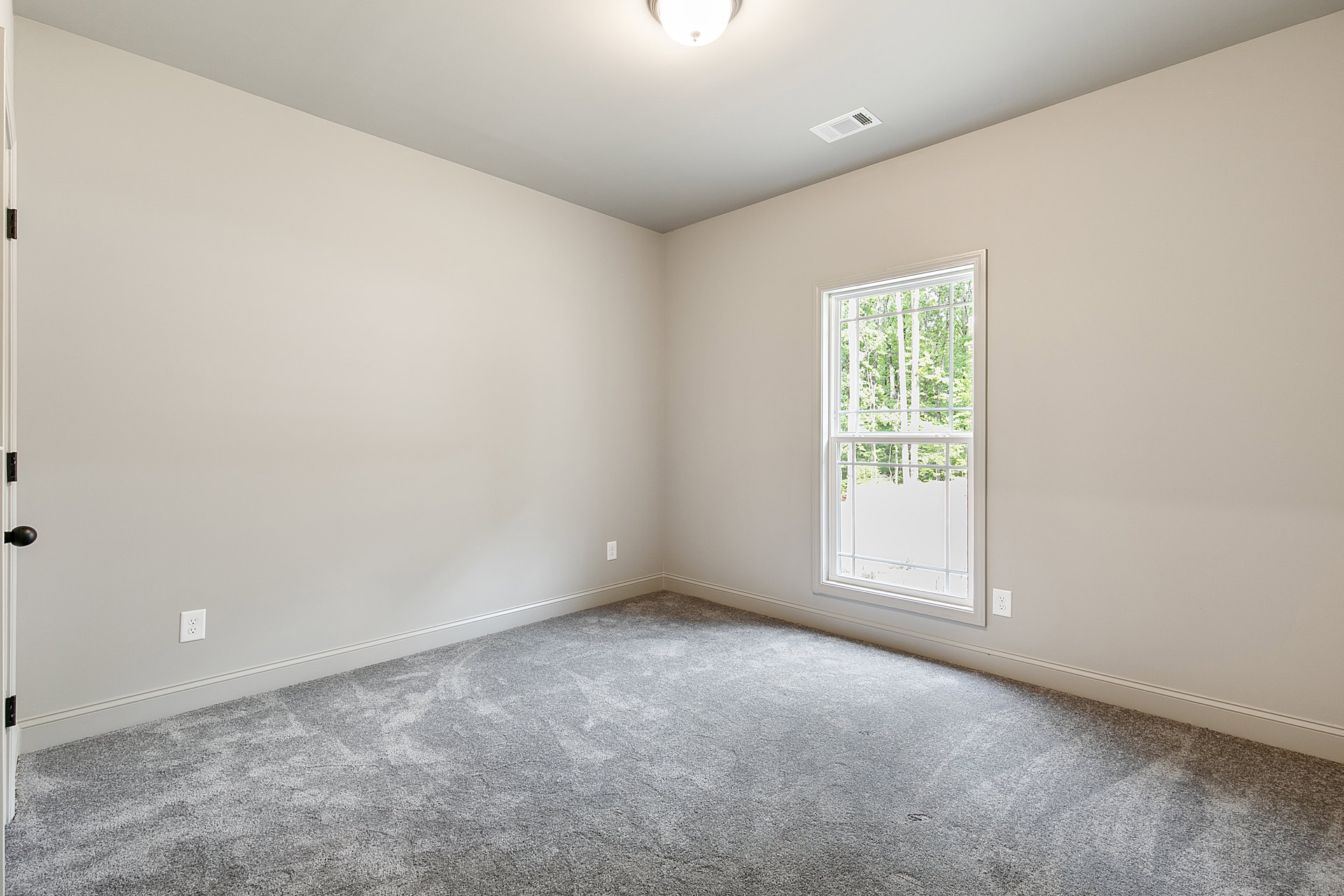 Sunlit room with grey carpet flooring, white walls, large window overlooking trees, white ceiling vent, and a partially visible door with a modern light bulb fixture.