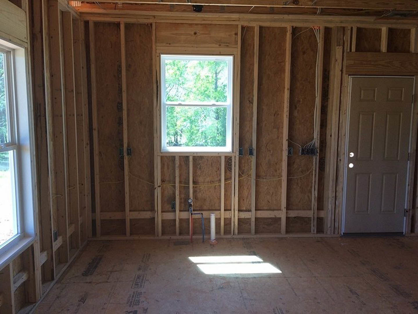 Sunlit room with wood plank flooring, large window framed in natural lumber, door with modern hardware, and recessed ceiling light illuminating the space.