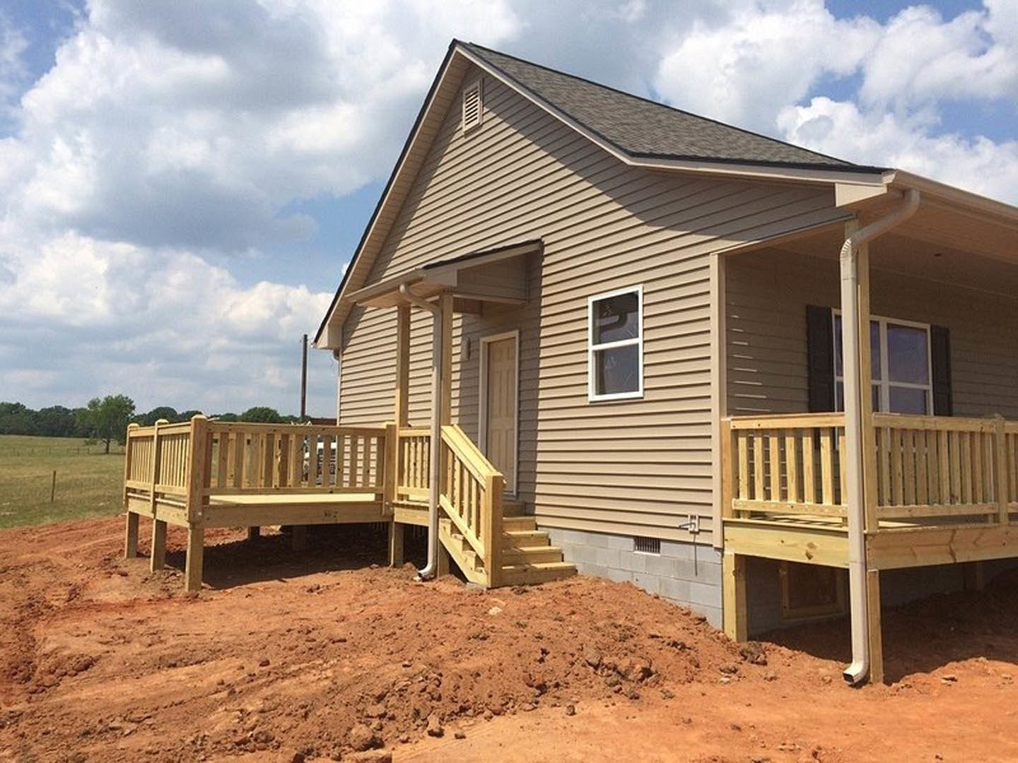 Wooden deck and stairs leading to a partially constructed house with white siding, white-framed window, white door with brown panel, and wooden bench on the ground.