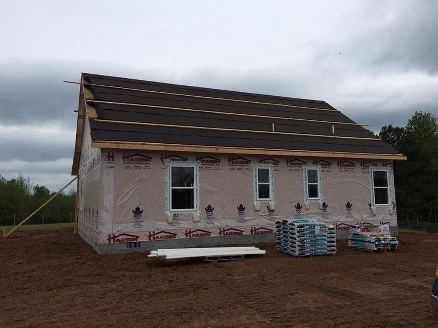 Two-story house framed with exposed wood and wrapped in plastic sheeting, pile of lumber and construction materials on dirt ground, cloudy sky and trees in background