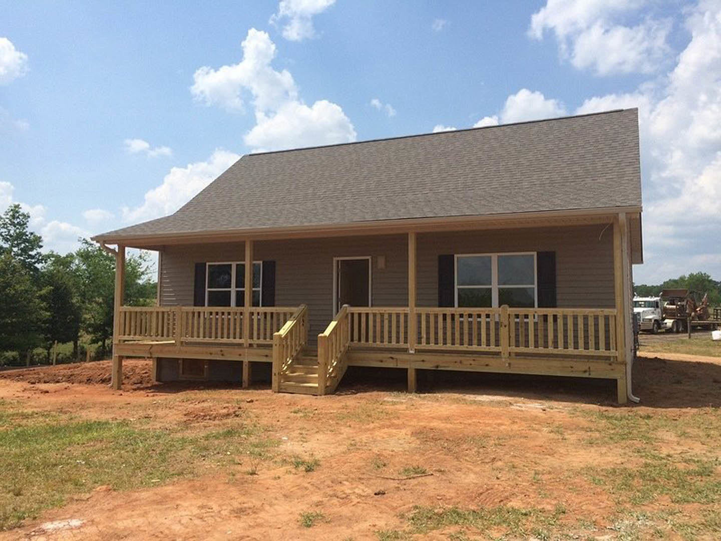 Two-story house with white siding, covered front porch with wooden railings and stairs, wooden deck, large windows, shingled roof, dirt and grass yard, bench near fence, trees and