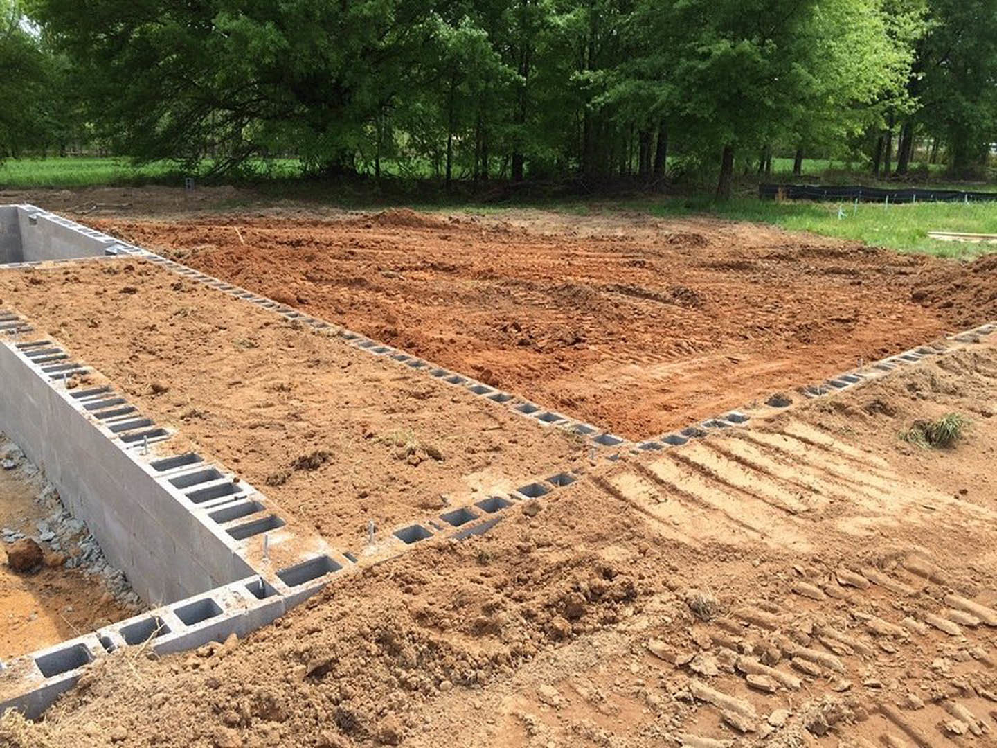 Concrete foundation slab with rectangular footings set in a dirt construction site, surrounded by grass and mature trees.