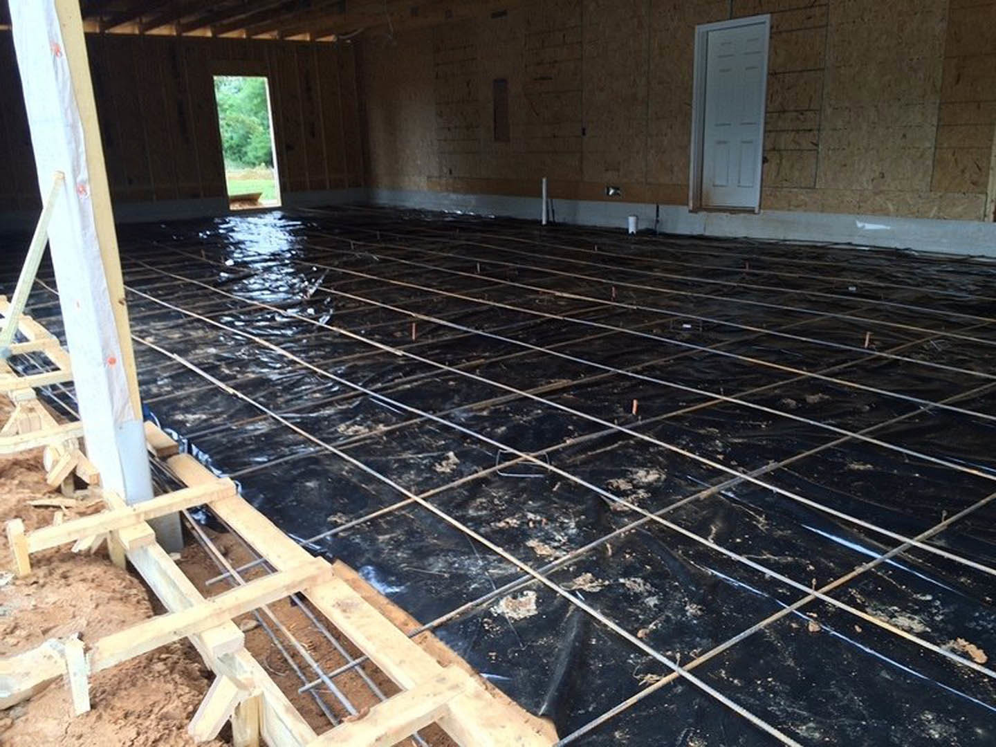 Freshly poured concrete floor with exposed metal rebar grid, white door with black trim, black tarp with white lines, and blurred forest visible through window