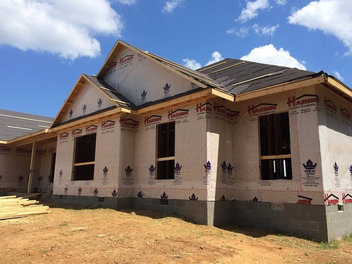 Framed house under construction with exposed wooden window frames, unfinished roof, and stacked lumber, set against a bright blue sky with scattered clouds