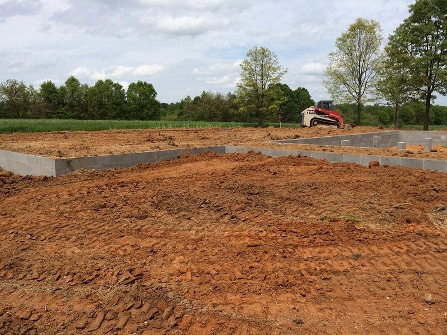 Bulldozer parked behind a square concrete foundation slab on a dirt construction site, surrounded by leafy trees under a cloudy sky